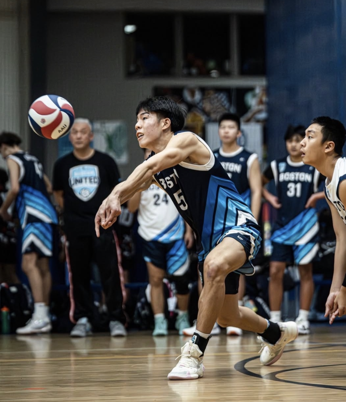 A male volleyball player in a blue and black uniform dives forward to hit a volleyball inside a gymnasium, with teammates and a coach watching in the background.