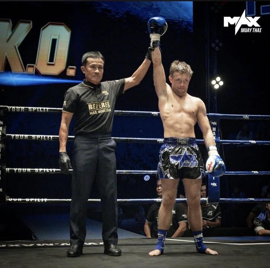 A Muay Thai fighter with a shirtless upper body and wearing shorts and gloves is having his hand raised by a referee in a boxing ring, indicating victory.
