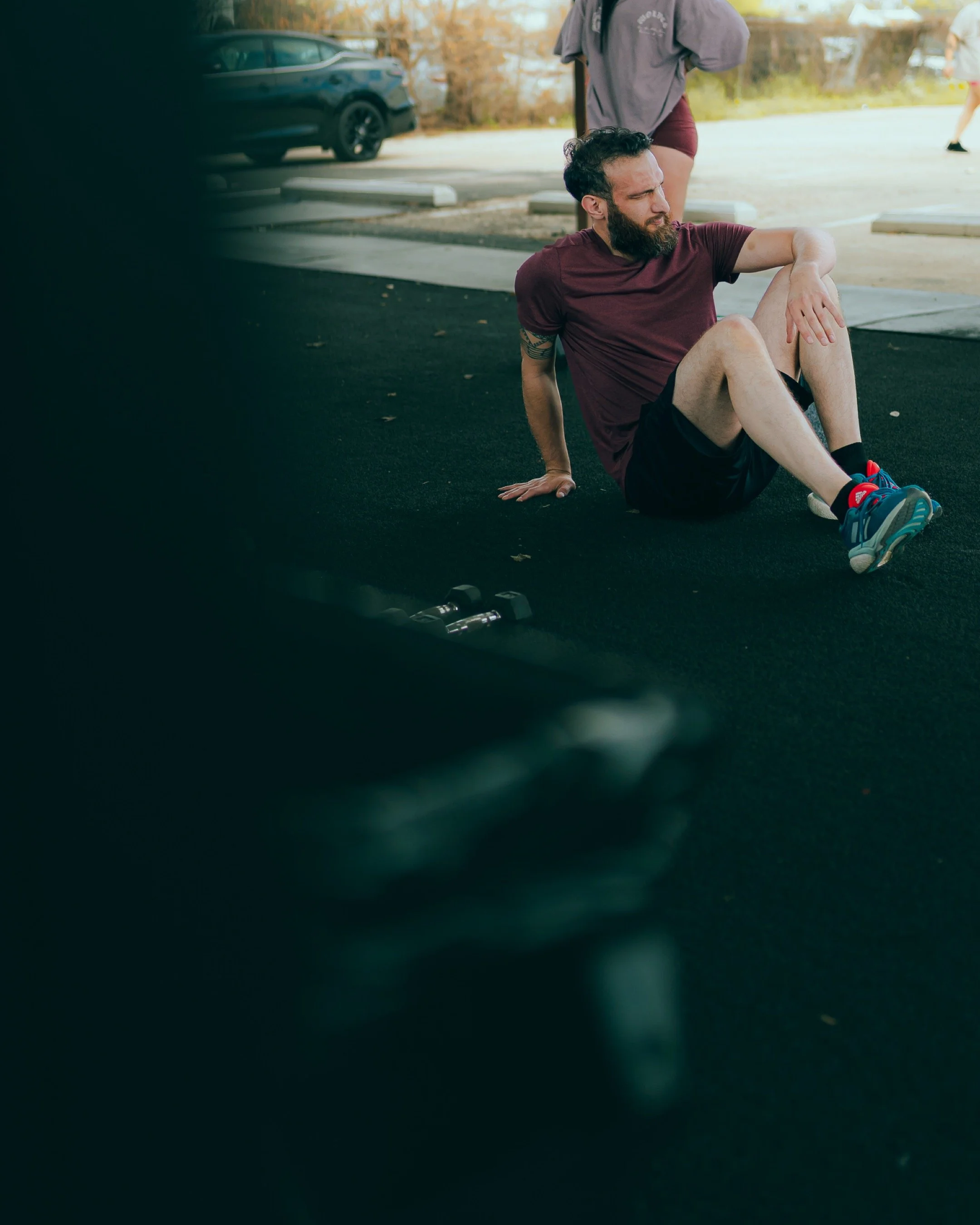 A man with a beard and tattoos on his arm sitting on a black gym mat outdoors, wearing a maroon T-shirt, black shorts, and blue athletic shoes, with another person standing behind him and cars in the background.