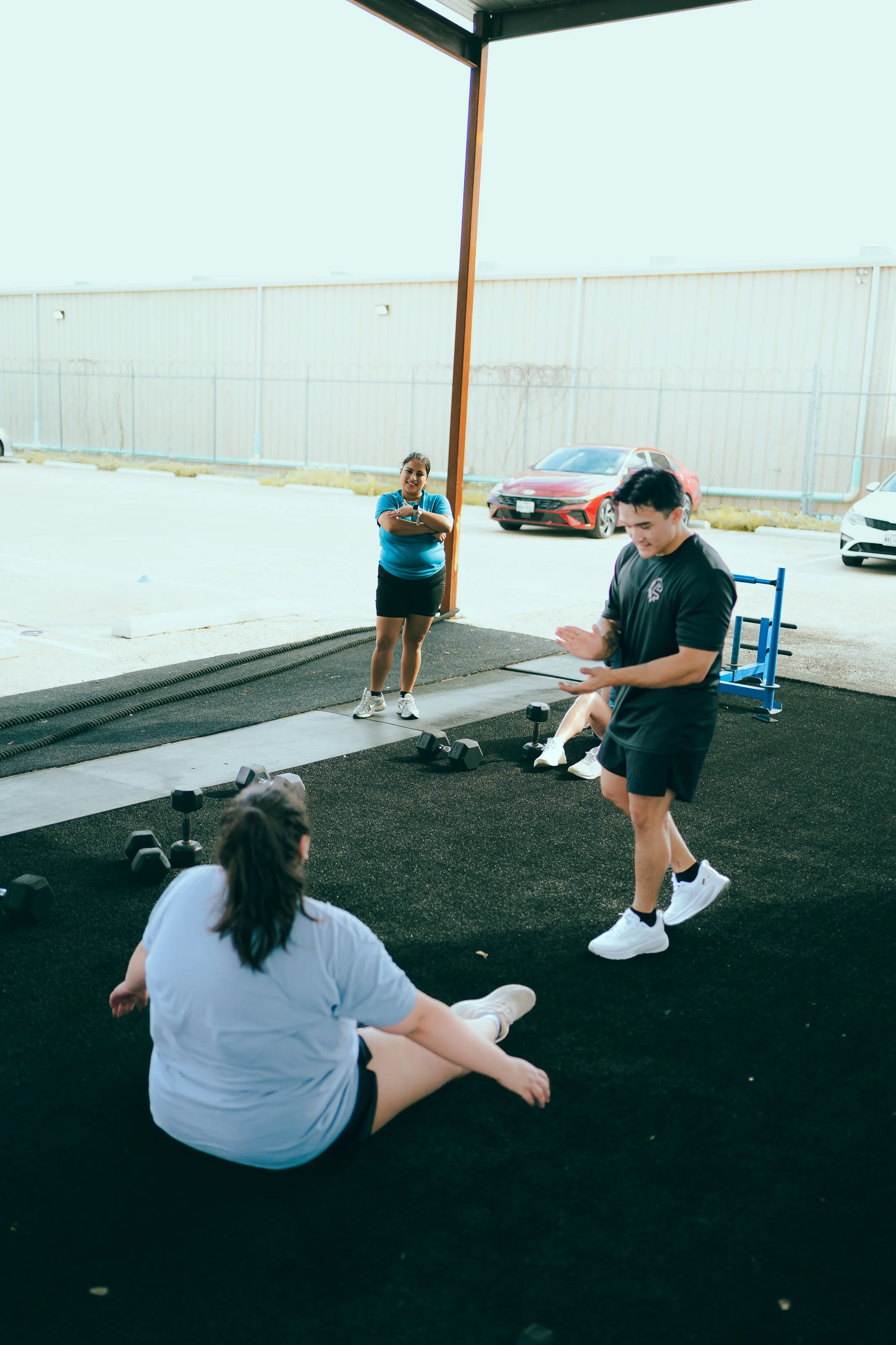 Group of young adults during workout session at outdoor gym in parking lot. One woman is standing with arms crossed smiling, another person is sitting on the ground stretching, and a young man is walking while looking down. Dumbbells are placed on the ground nearby.