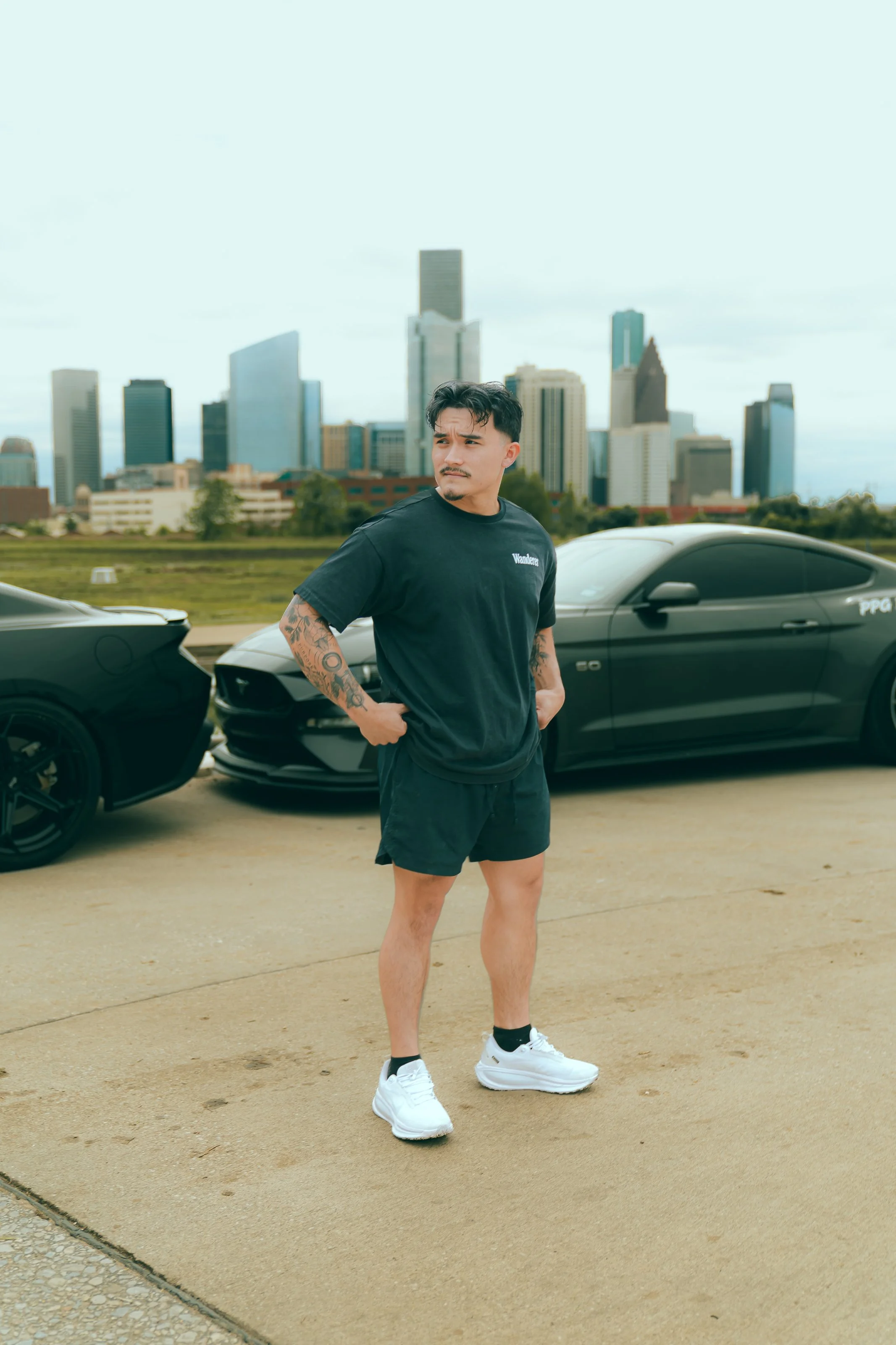 A man standing outdoors in front of a city skyline with tall buildings, wearing a black t-shirt, black shorts, white sneakers, and sporting tattoos on his arms, with two black sports cars parked behind him.
