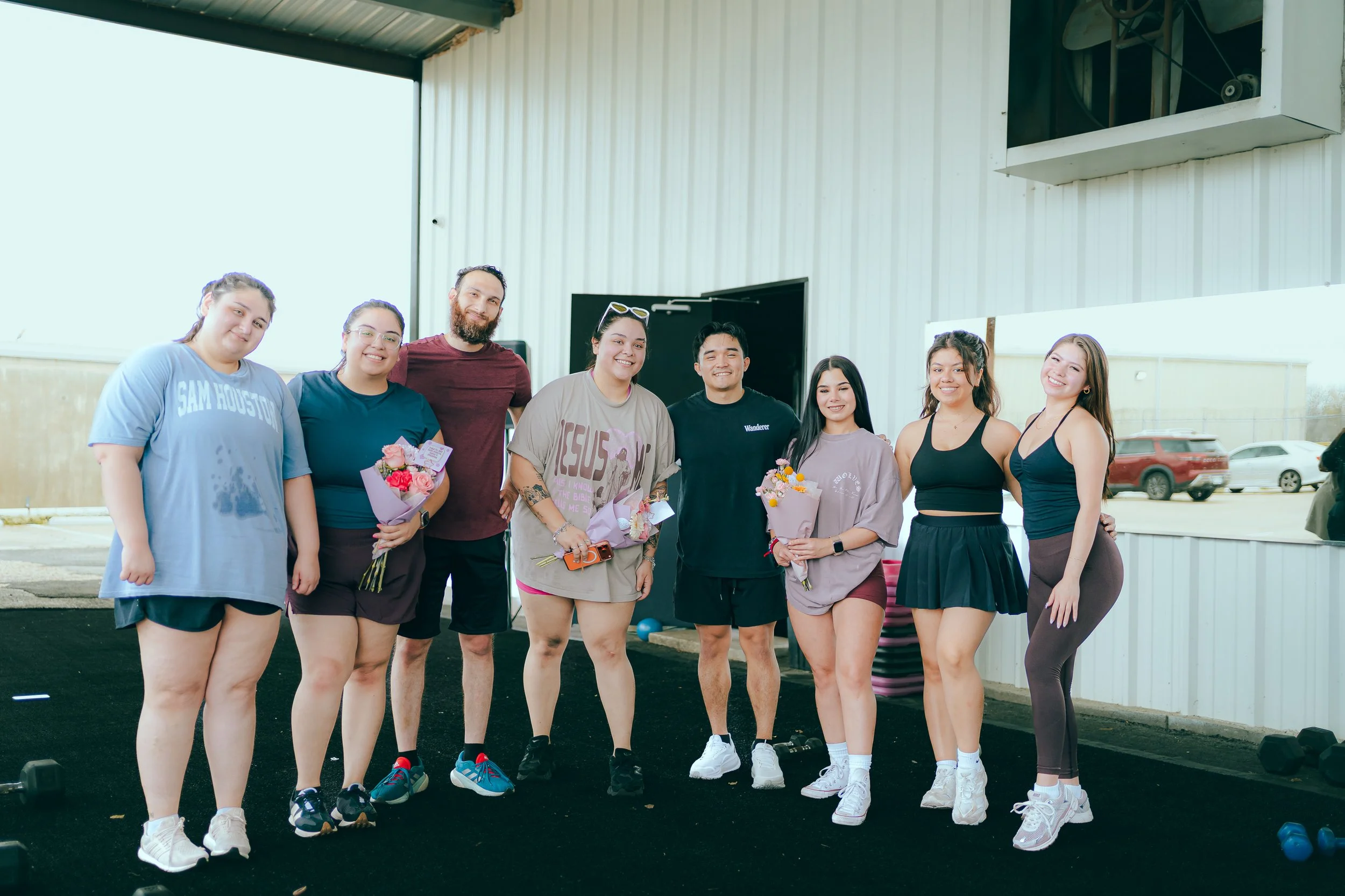 A group of nine diverse young adults standing outside, smiling, some holding bouquets of flowers, in front of a building with a metal roof. They are dressed casually and appear to be celebrating or gathering.