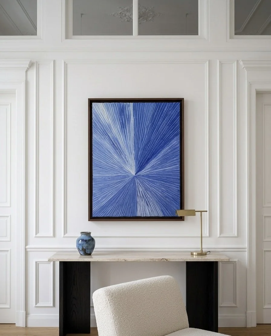 Ein modernes, minimalistisches Wohnzimmer mit weißer Wandverkleidung, einem blauen abstrakten Kunstwerk an der Wand, einer weißen Couch im Vordergrund, einer kleinen Vase auf einem Tisch und einer modernen Schreibtischlampe auf dem Tisch.