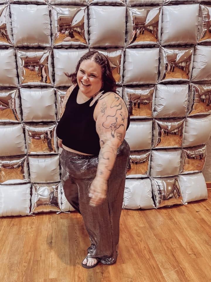 A woman with tattoos on her left arm, wearing a black sleeveless top and loose pants, smiling and pointing at the camera in front of a wall made of shiny silver foil balloons arranged in a grid pattern.