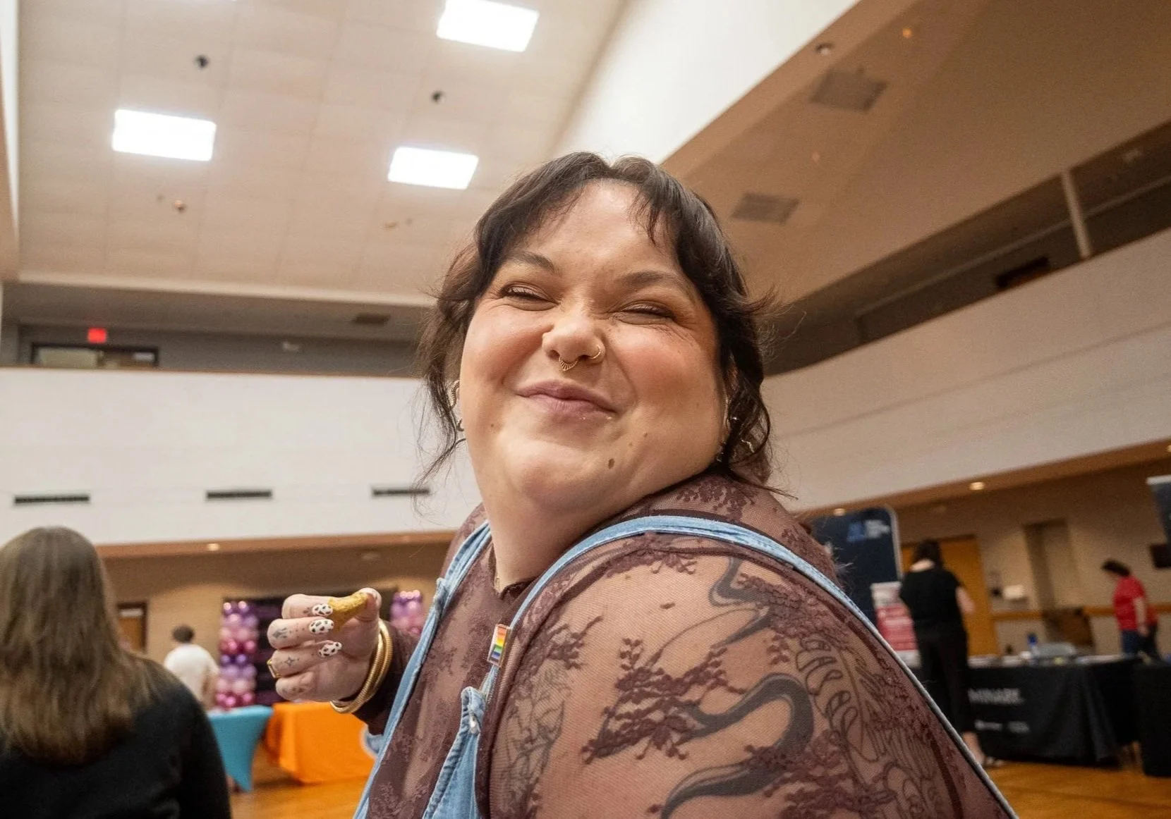 A woman with a septum piercing and tattoos, smiling and looking to the side in a large indoor event space with people, balloons, and tables in the background.