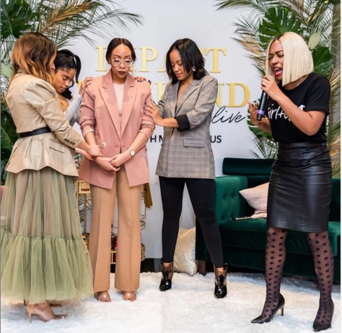 Five women standing close together on a white carpet, with a backdrop that reads "DIPAI CHAMP LIVE." The women appear to be praying or having a moment of reflection, with two women touching each other's shoulders. One woman on the right is holding a microphone and appears to be speaking or singing. They are dressed in fashionable attire, with the woman on the far right wearing a black leather skirt, polka dot tights, and high heels, and the woman second from the left wearing a pink pantsuit.