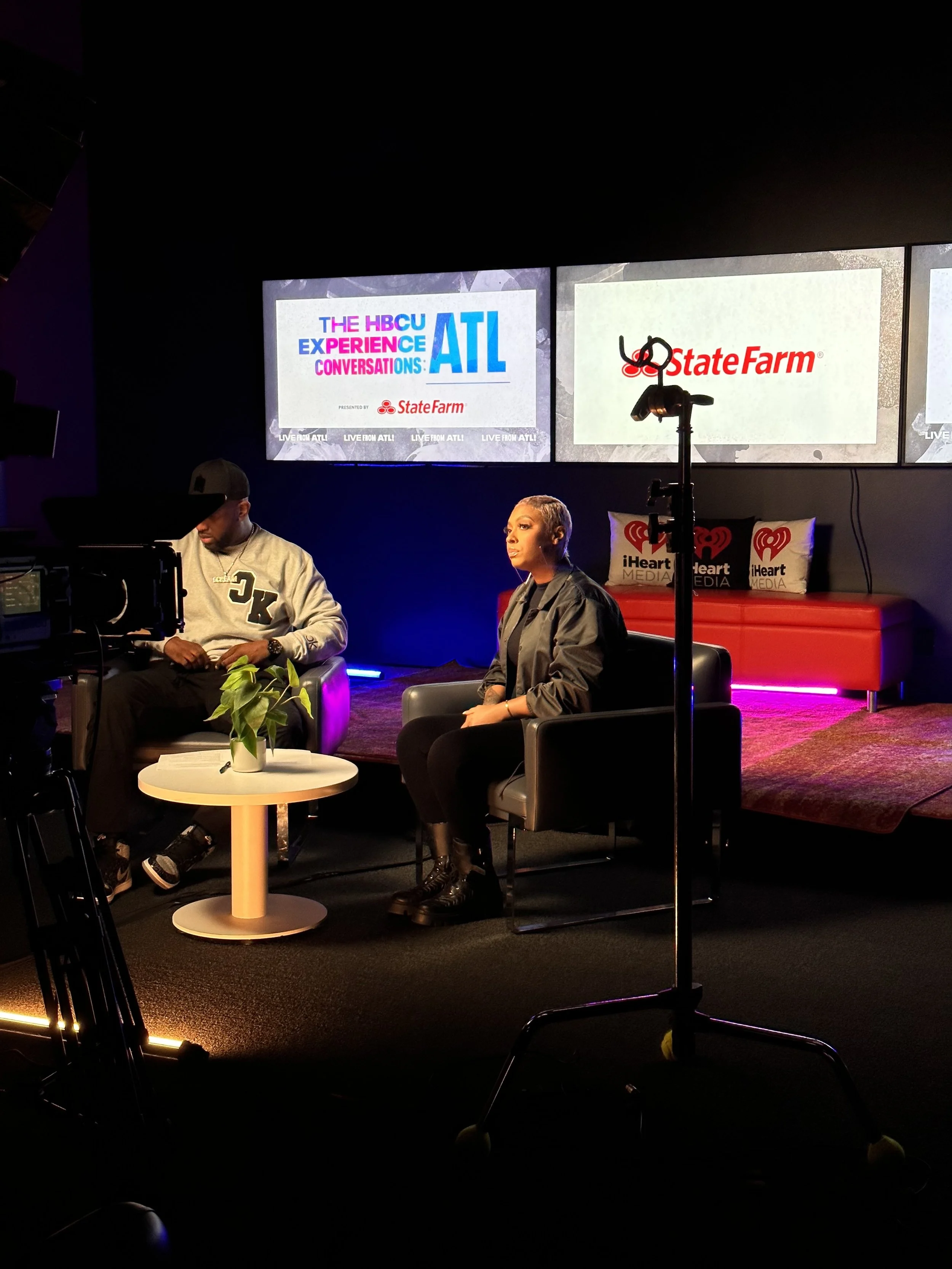 Studio setting with two people seated; a man on the left wearing a beige sweatshirt and cap, and a woman on the right with short blond hair and a black jacket. Behind them are three screens displaying event promotions with the logo of iHeartMedia and State Farm.