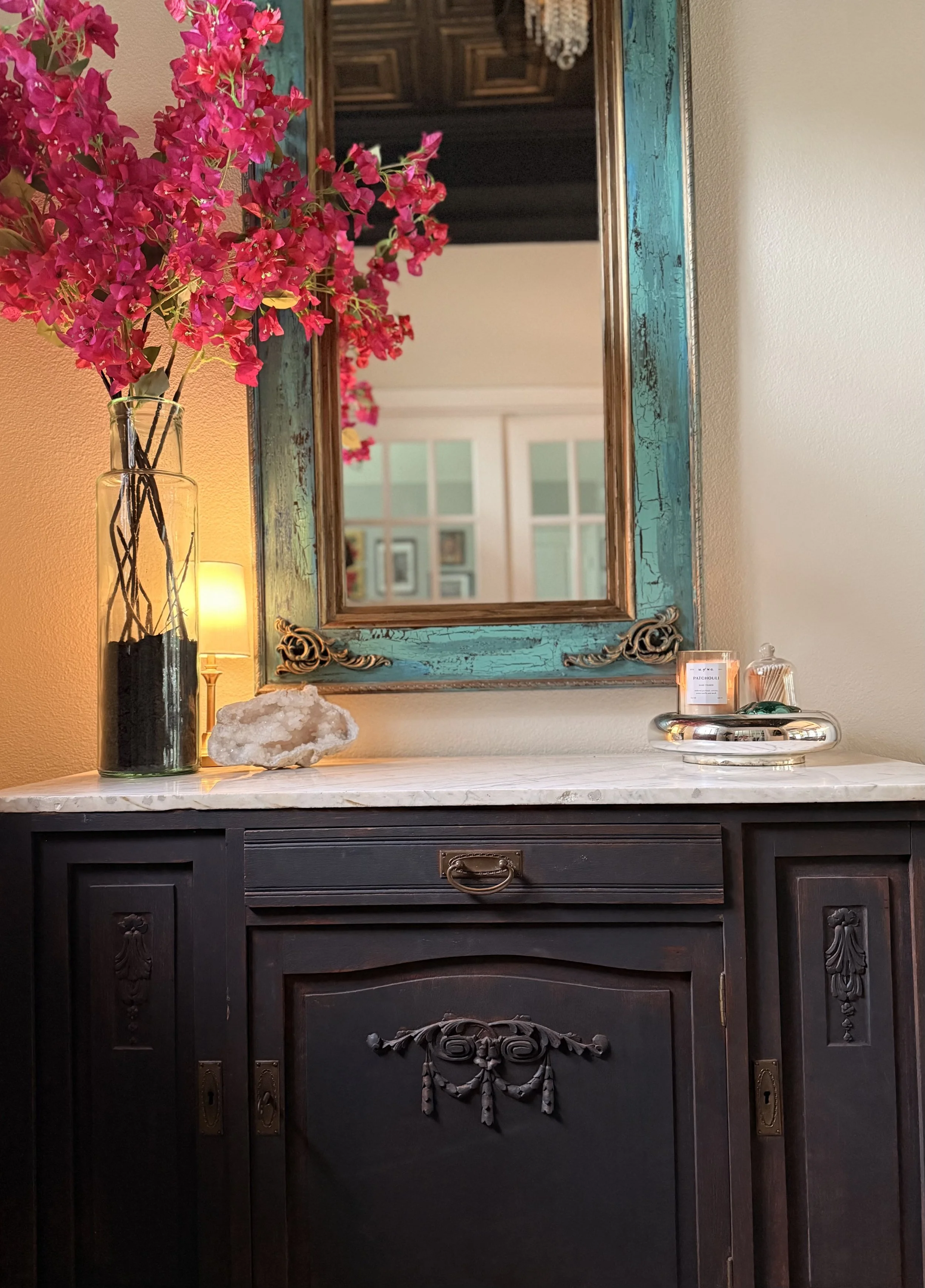 A vintage black wooden dresser with ornate carved details, a white marble top, and a large mirror with a distressed turquoise wooden frame and gold accents. On the dresser, there is a tall glass vase with pink bougainvillea flowers, a large white geode, a candle, and some small decorative items.