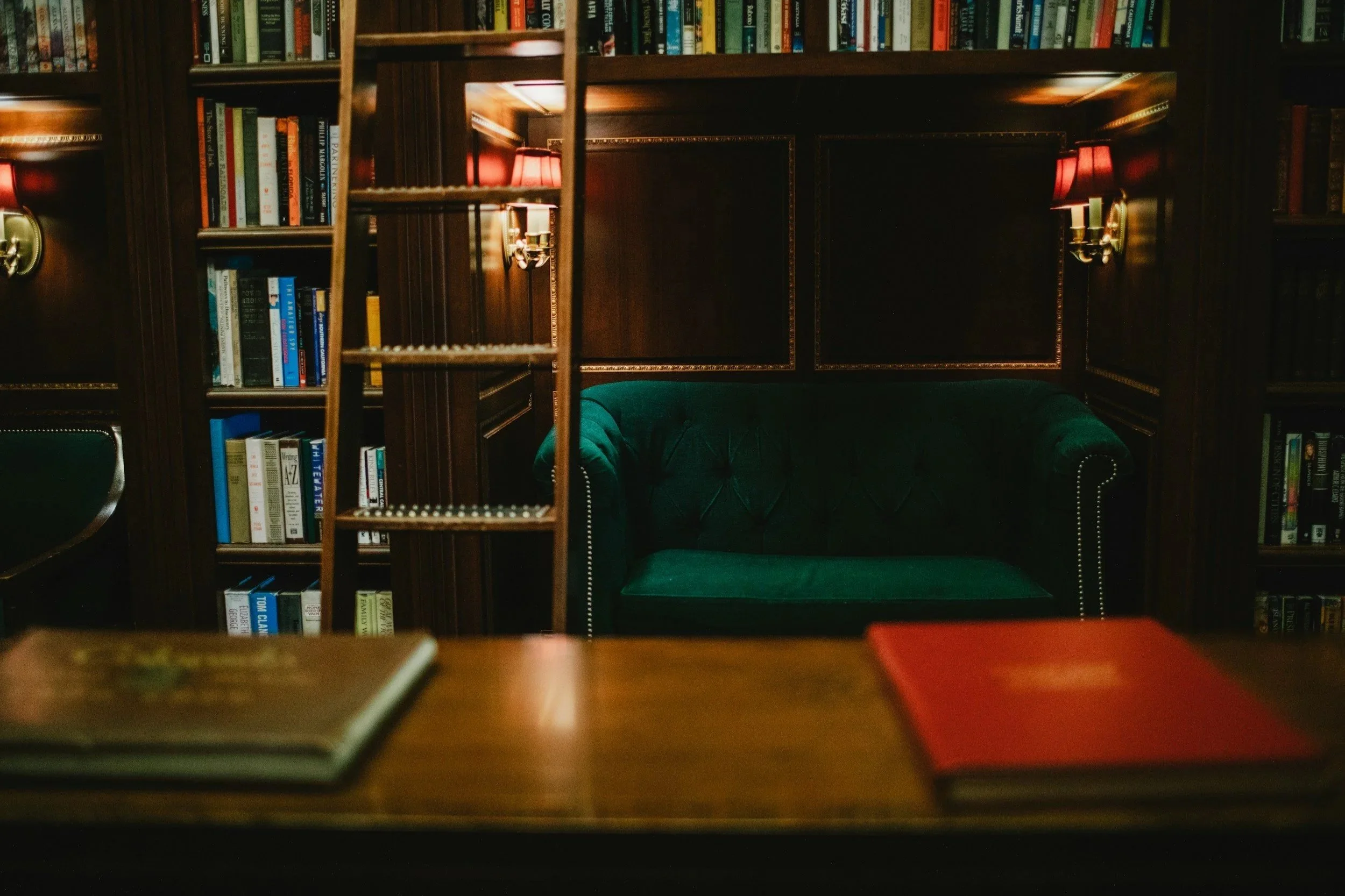 View of a cozy library room with dark wooden shelves filled with colorful books, red wall lamps, a green upholstered armchair, and a ladder for reaching high shelves.