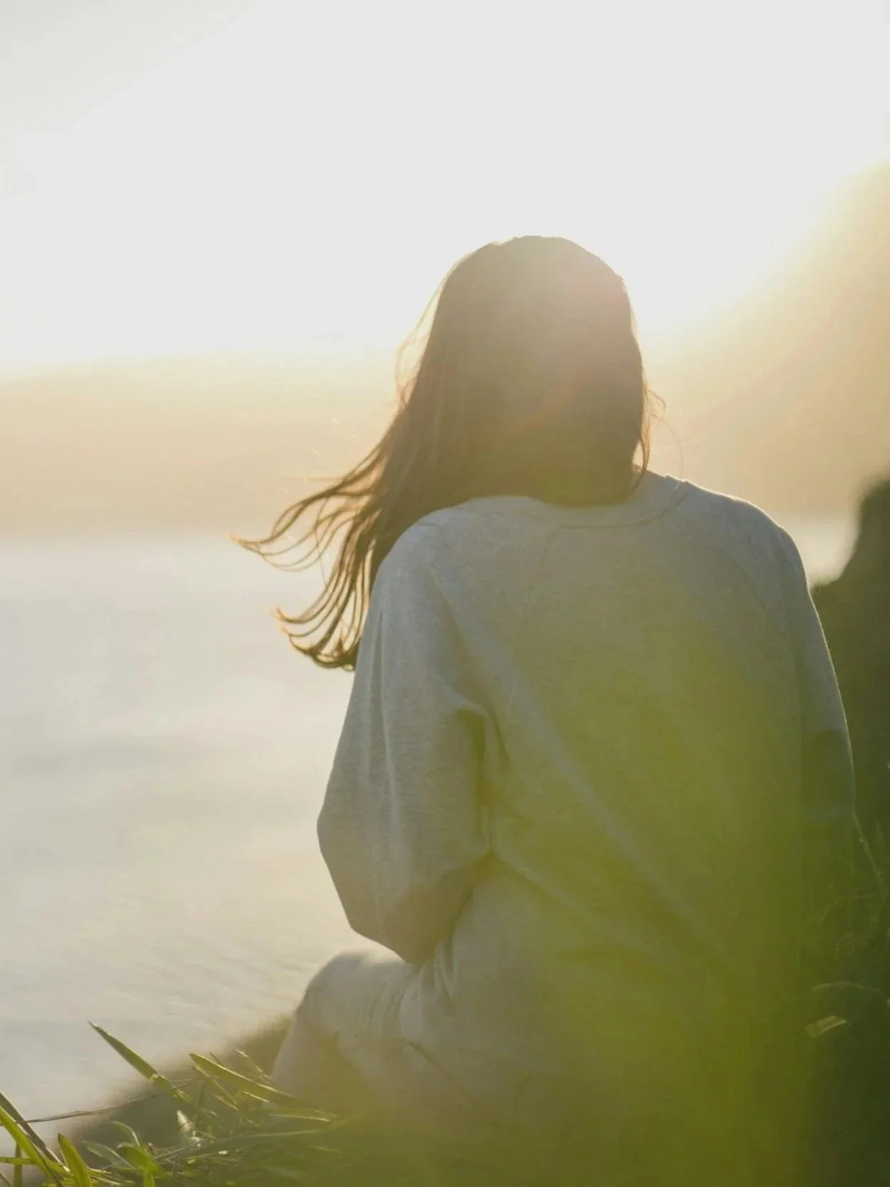 A person with long hair, wearing a grey hoodie, is sitting outdoors near water, facing away from the camera, with the bright sun in the background.