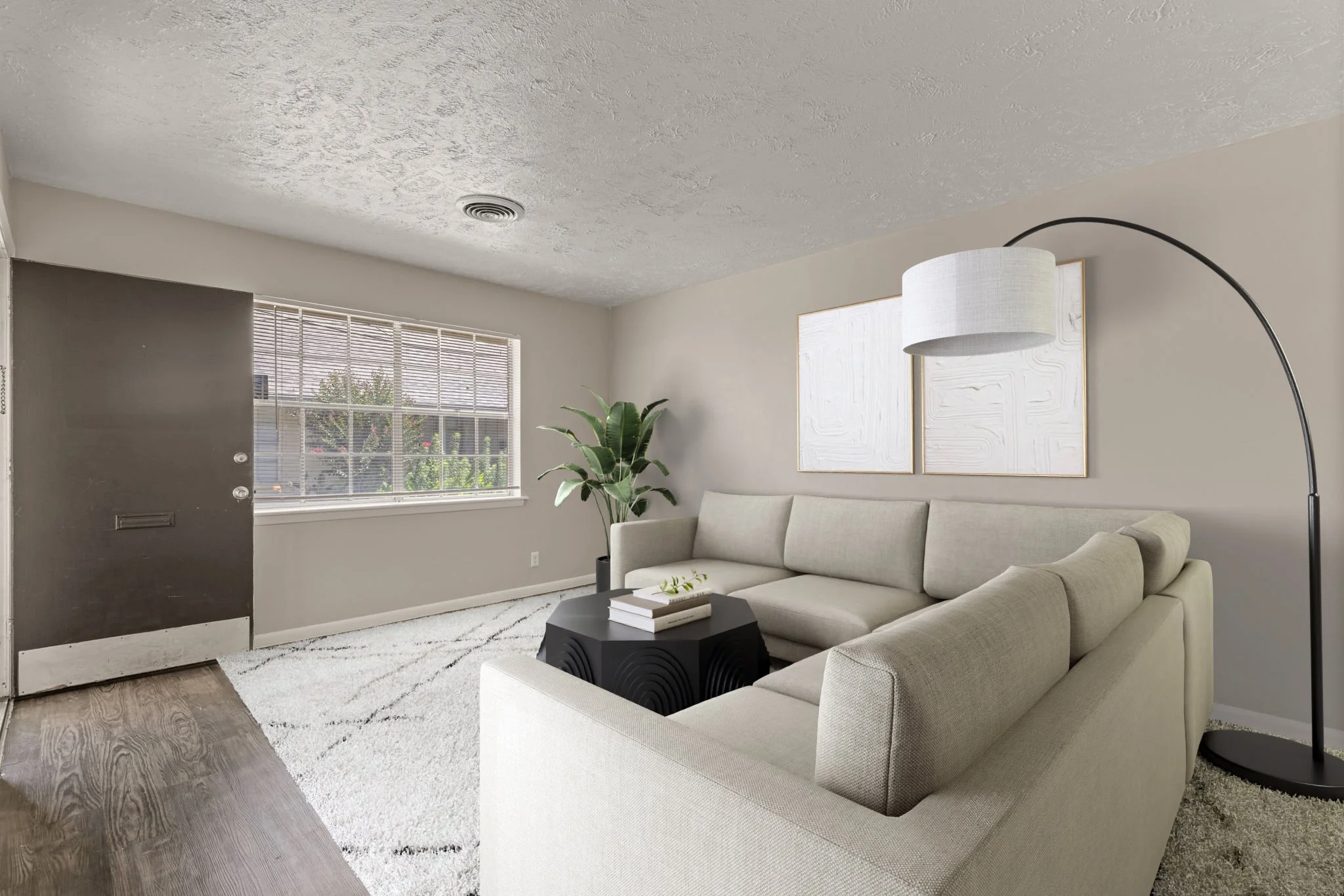 A living room with a beige sofa, a black coffee table with books, a large white floor lamp, a potted plant, a textured white wall art, window with blinds, and a window view of trees outside.