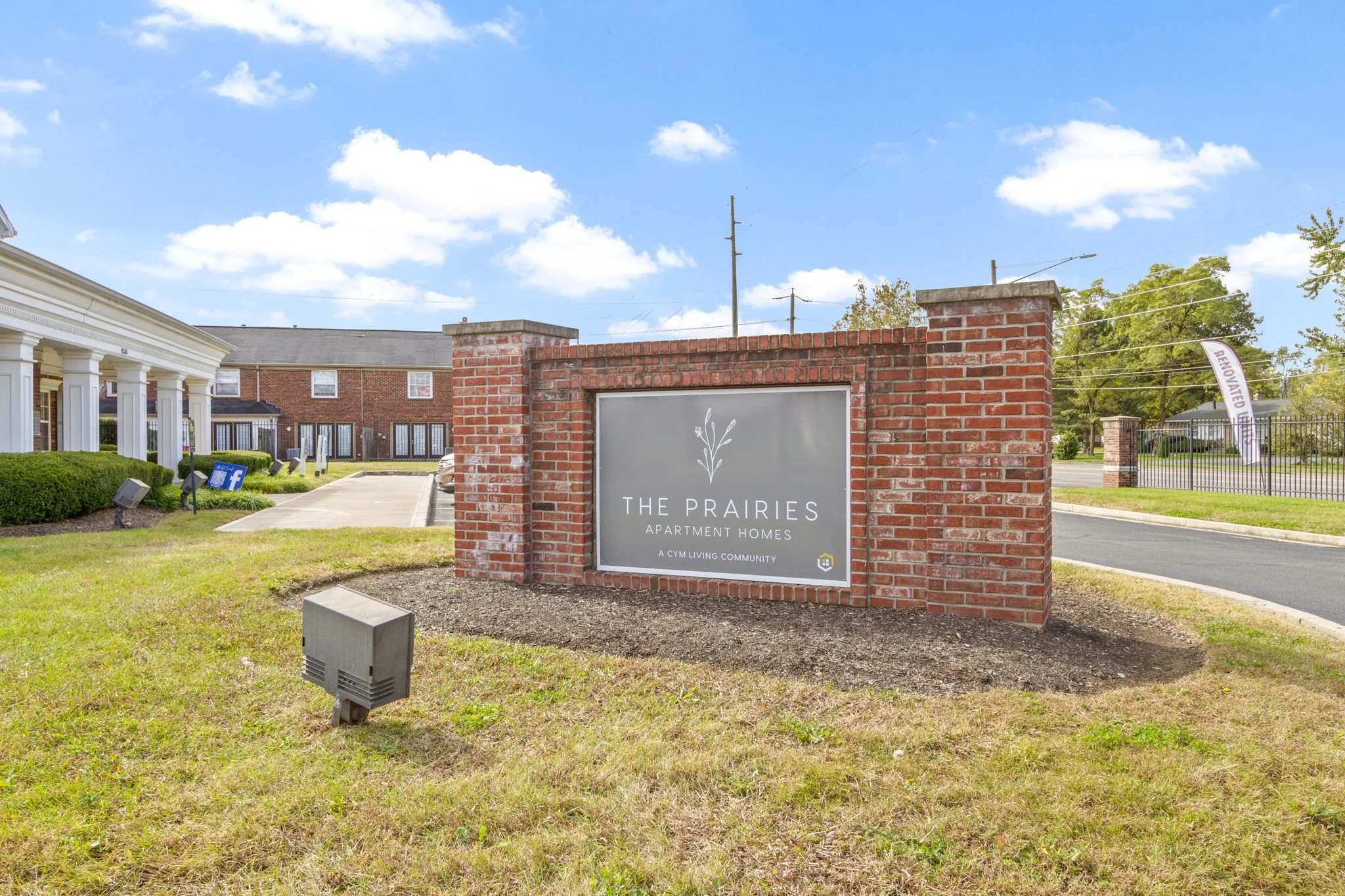 Sign for The Prairies apartment homes on a brick wall at entrance of the community, with a background of buildings, fence, and trees under a partly cloudy sky.