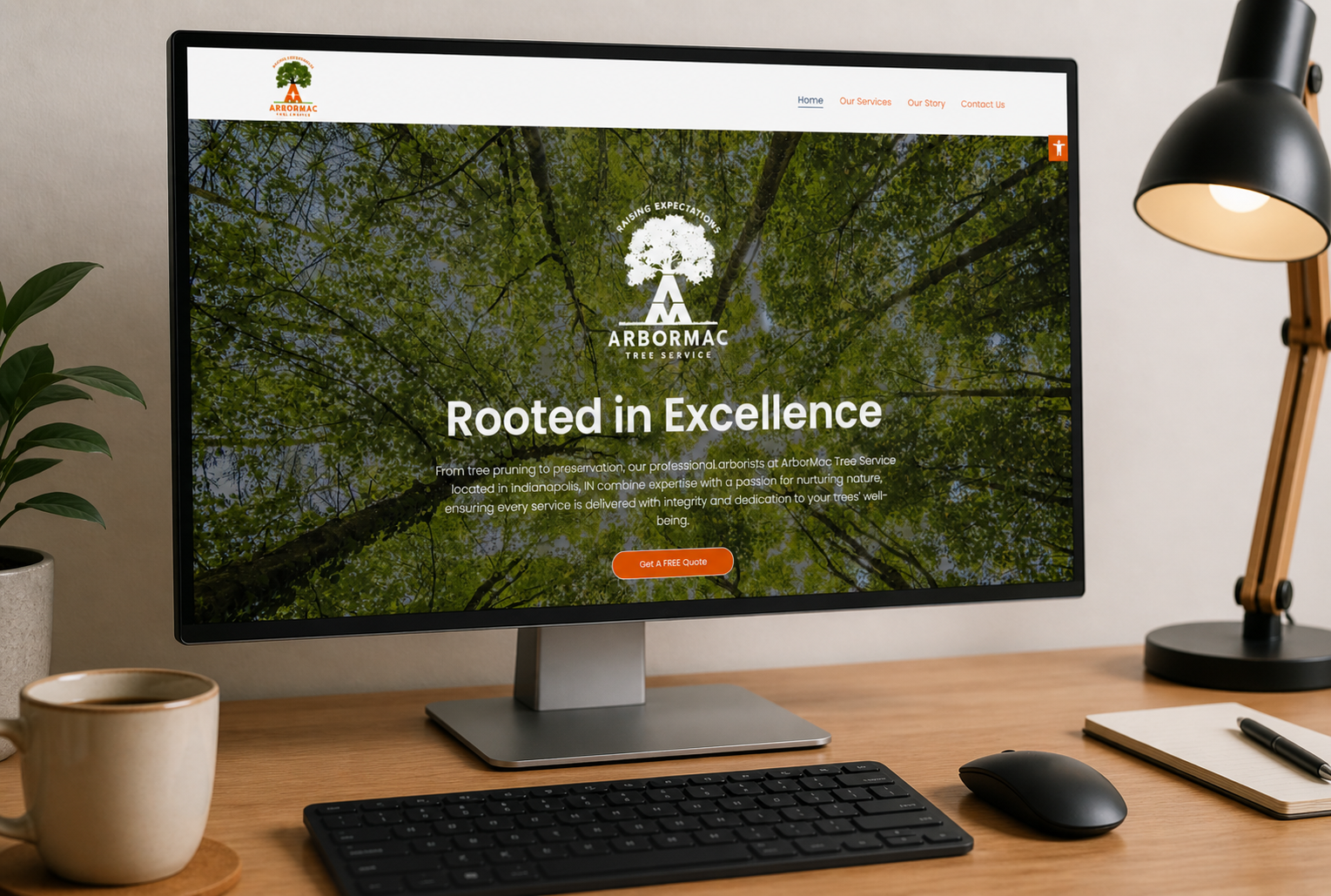 Computer monitor displaying Arbormac Tree Service website with a background image of trees and leaves, on a wooden desk with a black keyboard, mouse, notepad with a pen, a coffee mug, a small plant on the left, and a desk lamp on the right.