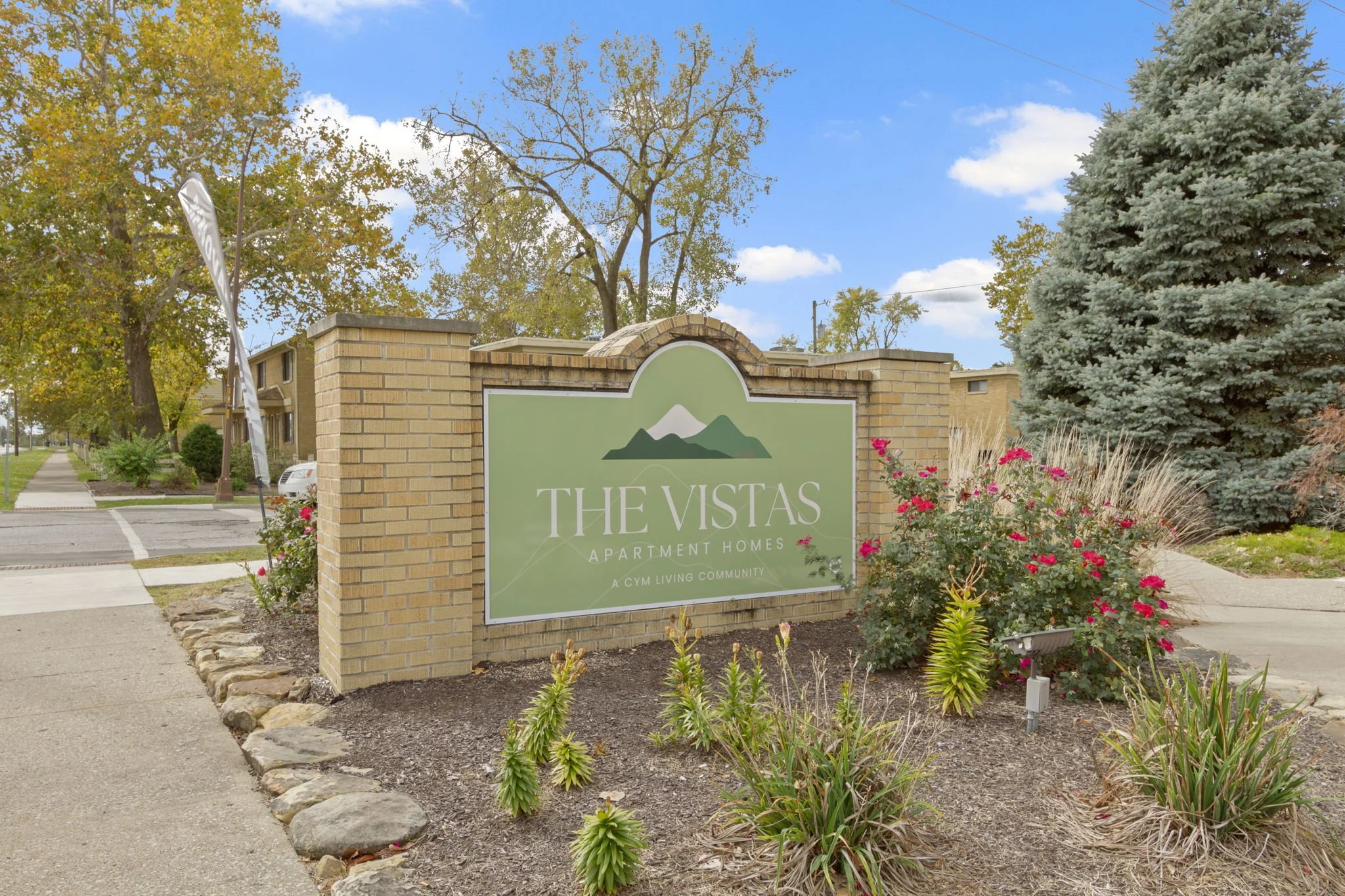 Entrance sign for The Vistas apartment homes in a landscaped area with flowers and trees, under a blue sky with clouds.