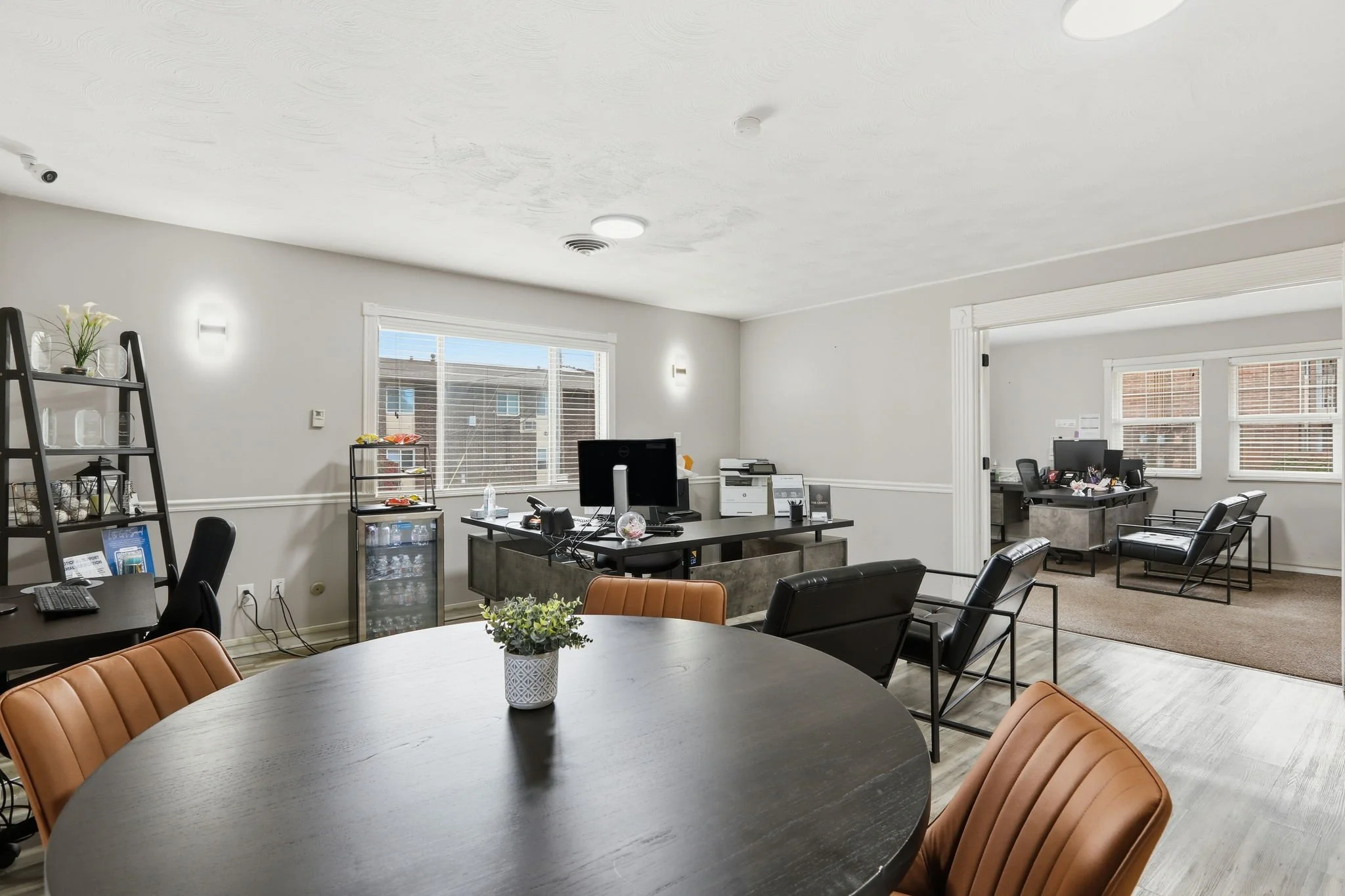 An office with a round table in the foreground, surrounded by brown chairs, and multiple desks and chairs in the background. Windows let in natural light, and there are various office supplies and decorations visible.