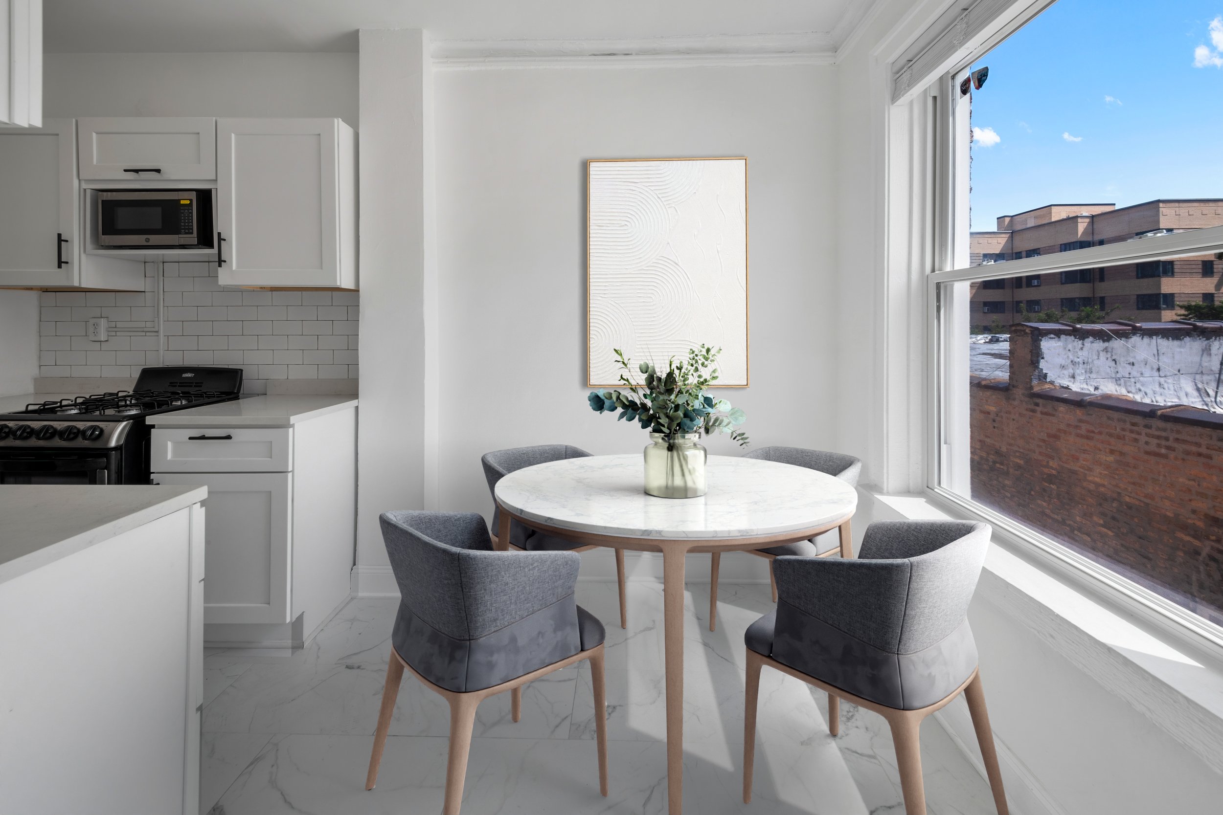 Bright dining area with a round marble table, four gray upholstered chairs, a glass vase with greenery, and a large window showing a city view.