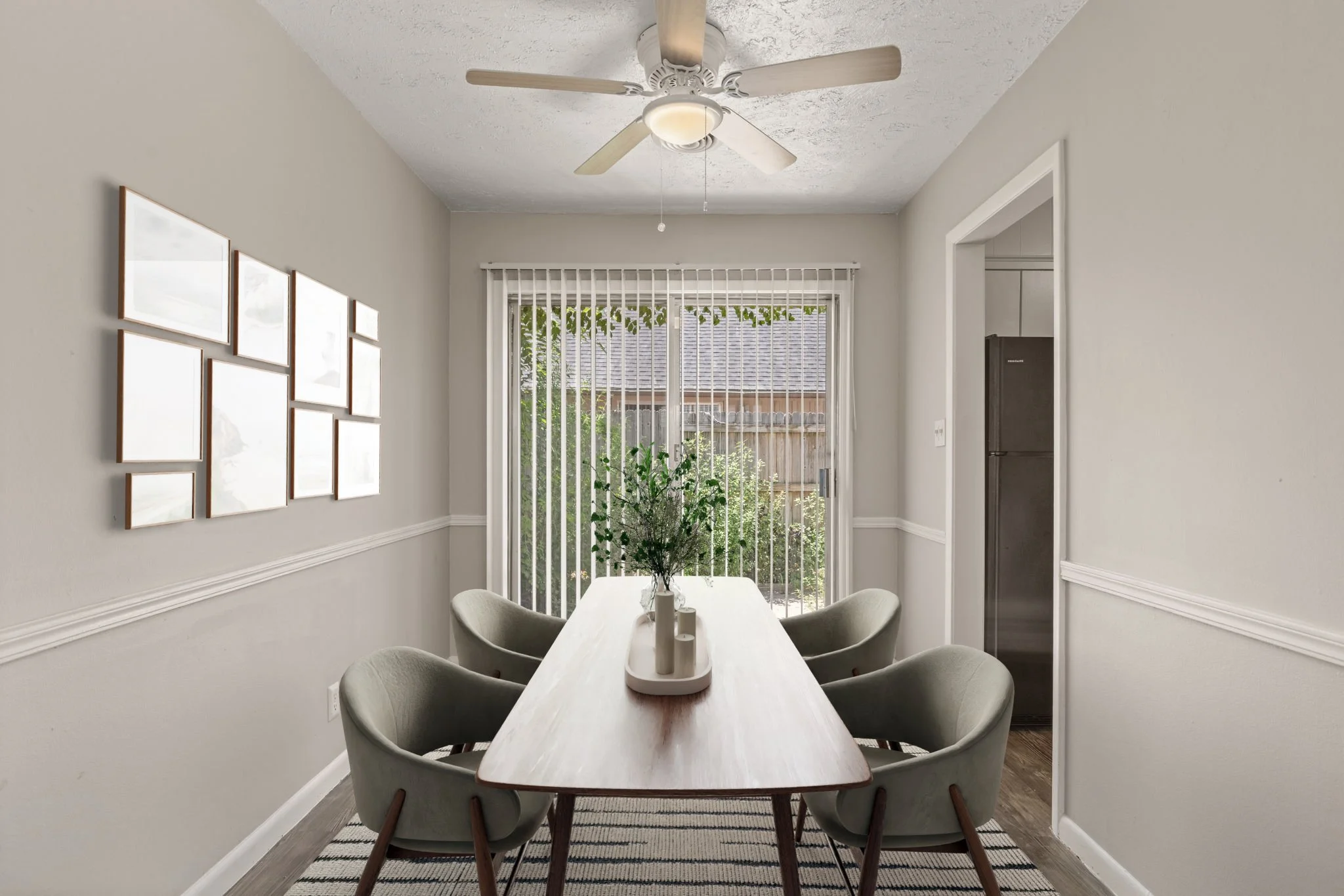 A dining room with a rectangular wooden table surrounded by six light-colored upholstered chairs. A small vase with green plants and three candles sit on the table. Natural light enters through sliding glass doors with vertical blinds, leading to a b