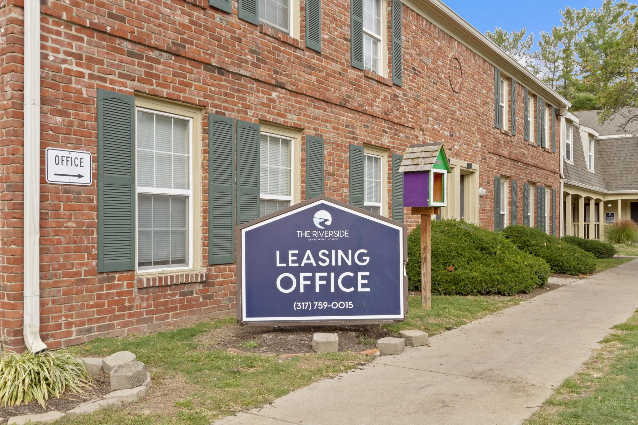 Red brick apartment building with green window shutters and a large blue leasing office sign in front, advertising leasing office contact information, and a colorful birdhouse on a wooden post nearby.