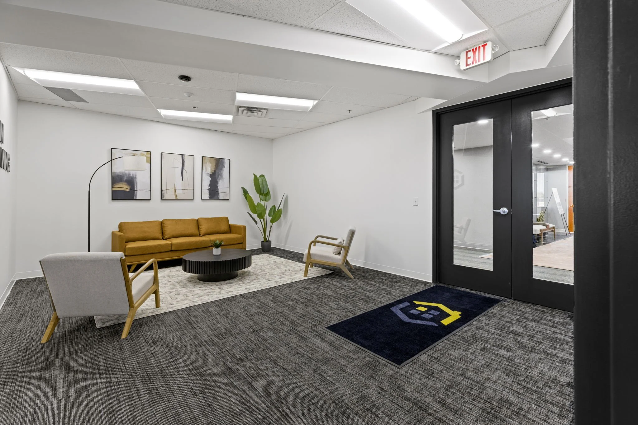 A modern office waiting area with a mustard-colored sofa, two chairs, a small round coffee table, abstract art on the wall, a tall green plant, and a dark entrance door with an exit sign above.
