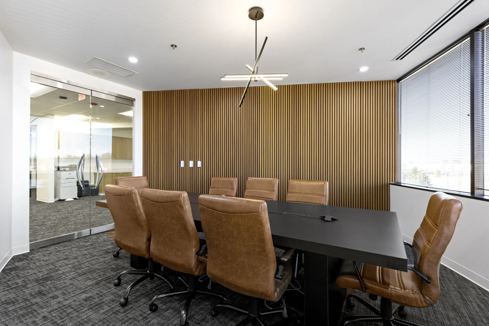 A modern conference room with a long black table surrounded by brown leather chairs. There is a wooden slat accent wall and large windows with blinds, plus contemporary ceiling lights.