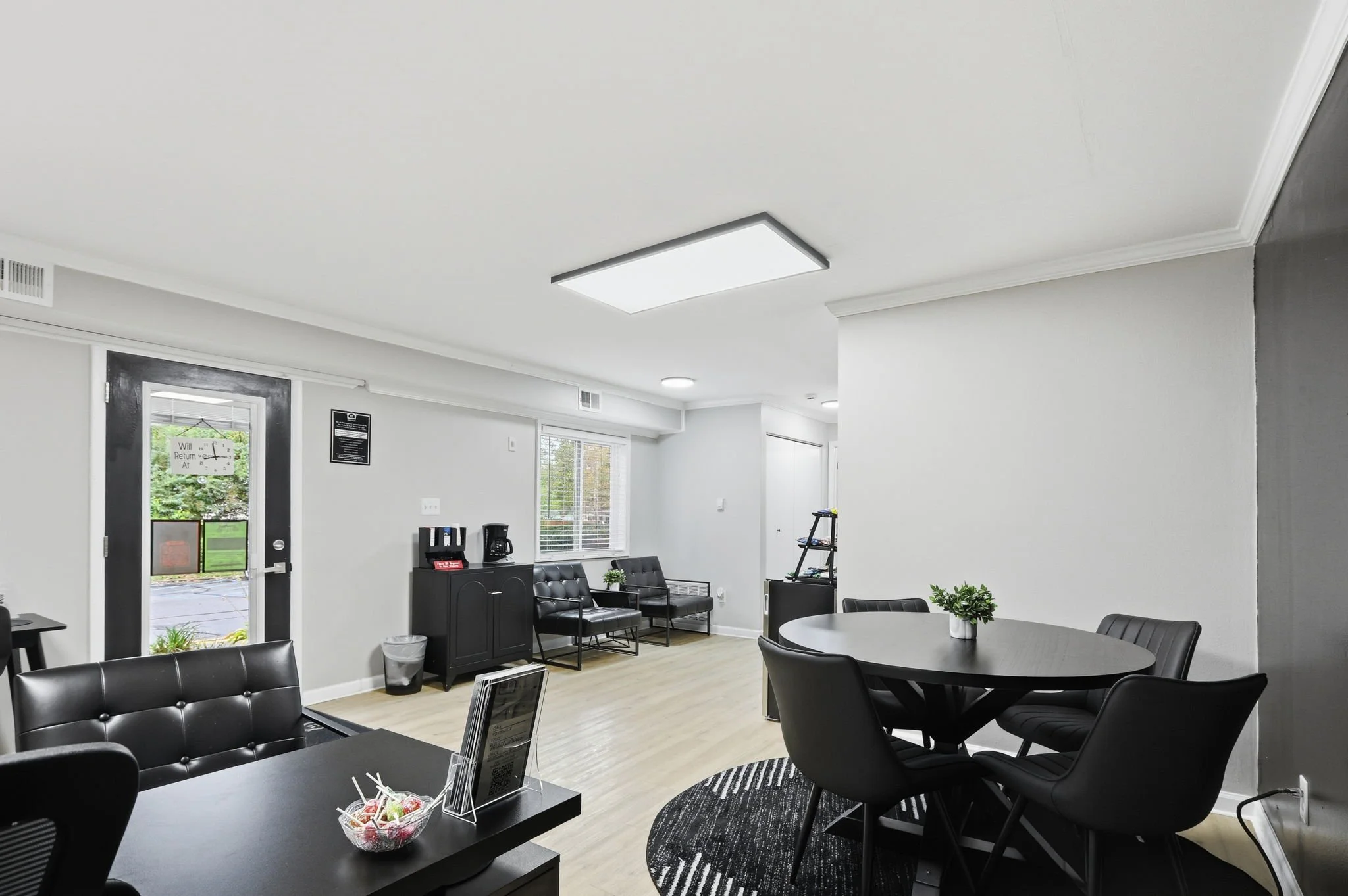 Interior of a modern waiting or lounge area with black chairs and a round table, white walls, large windows, and ceiling lights.