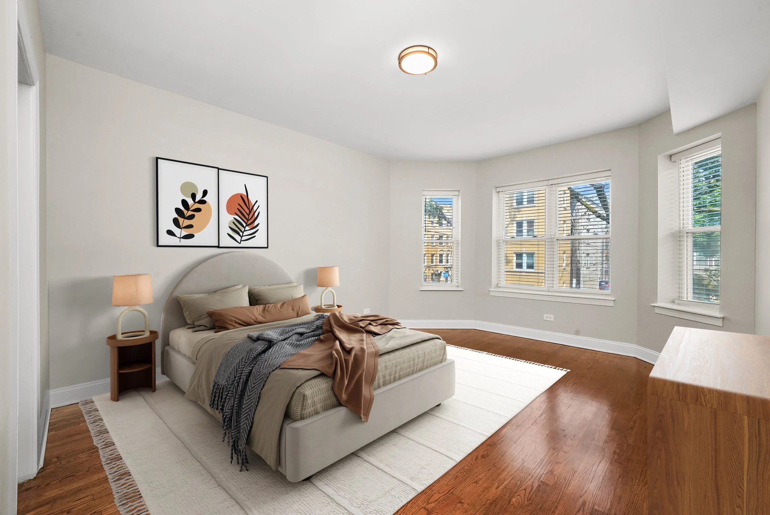 Bright bedroom with a white bed, beige headboard, and bedding. Two nightstands with matching lamps on either side. Two framed abstract art pieces above the bed. Large windows with blinds showing buildings outside. Wooden floor and white walls.