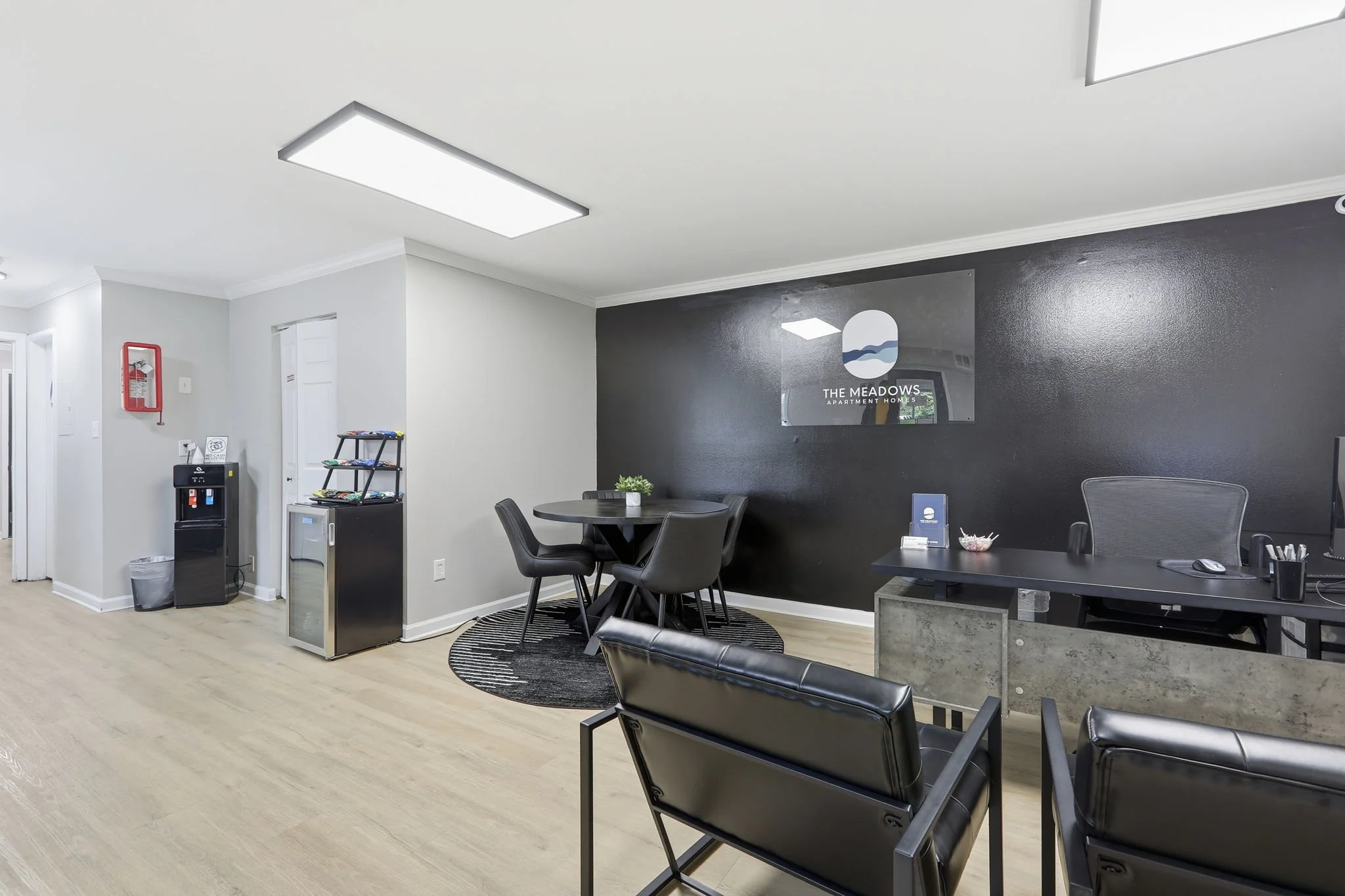 Interior view of an office lobby with black accent wall, round black rug, black chairs, a table, a sign for The Meadows Apartment Homes, and office desk with supplies.