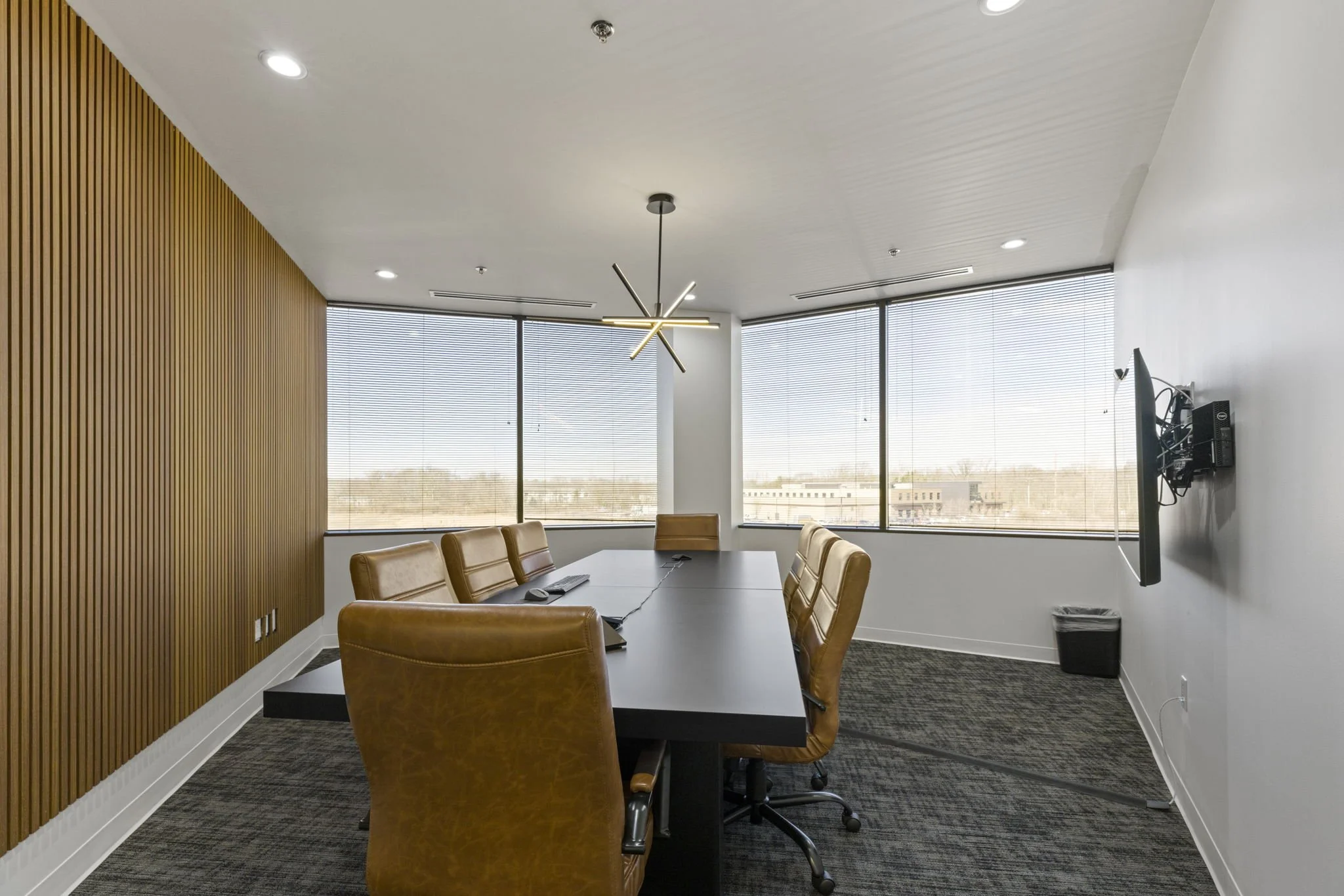 Modern conference room with a long black table, tan leather chairs, wooden accent wall, large windows with blinds, and a decorative ceiling light fixture.