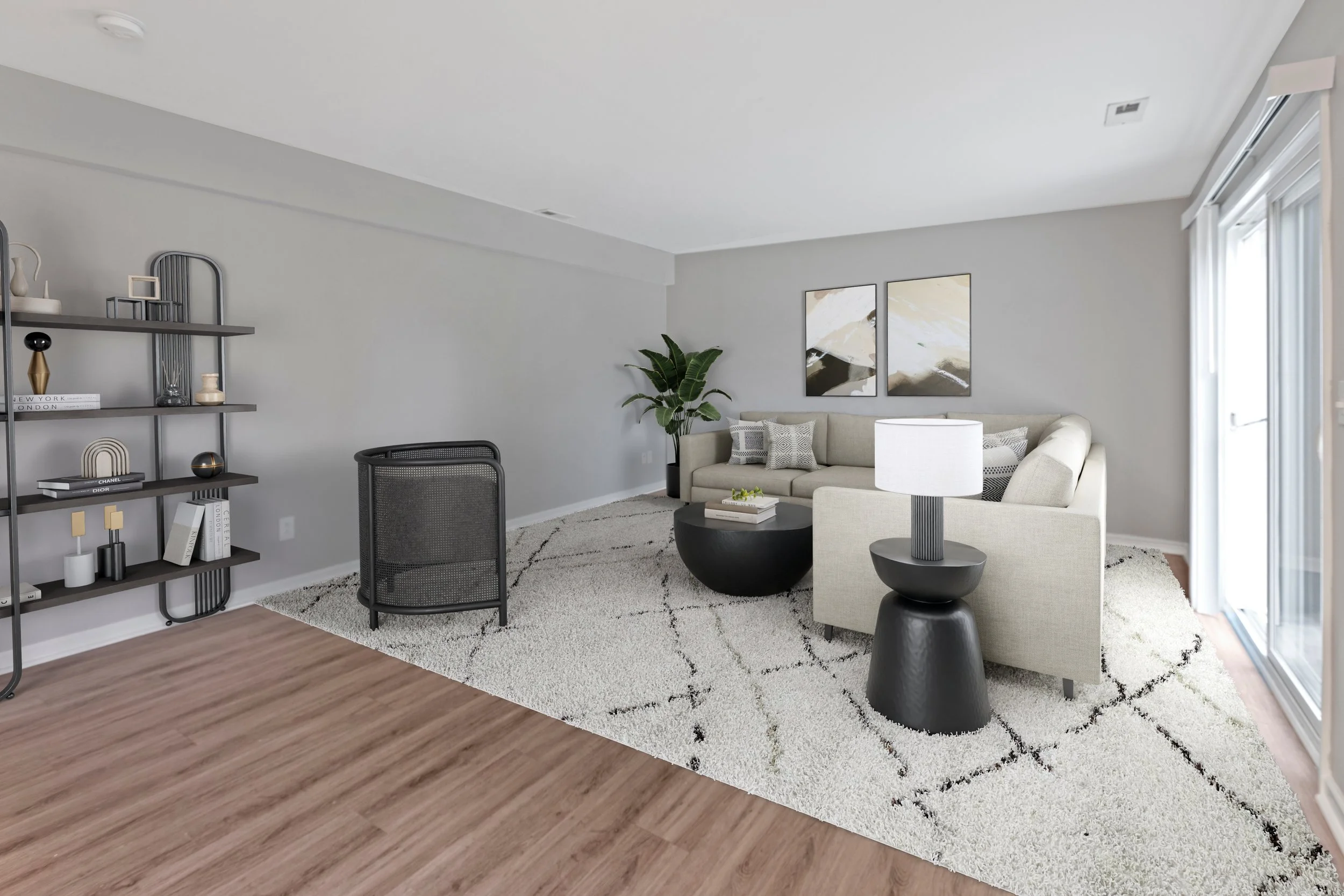 A living room with a beige sectional sofa, abstract wall art, a tall green plant, a coffee table, and a side table with a white lamp. A black armchair, a gray bookshelf with decorative items, and a large window with white blinds are also visible. The