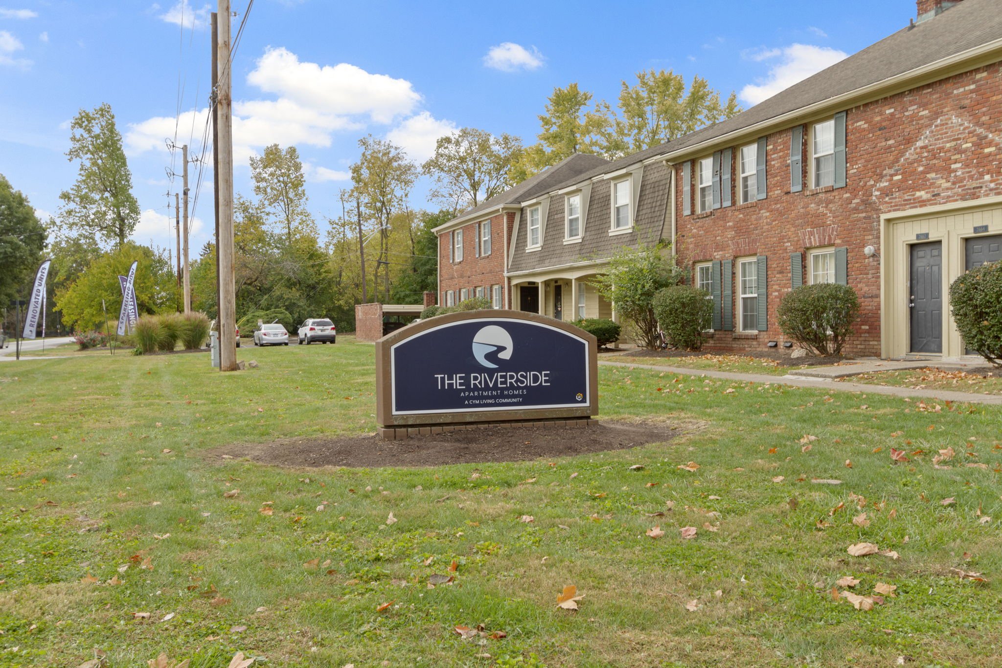 A sign in the front yard of a residential apartment complex named 'The Riverside' with a logo of a river and the tagline 'A CYM Living Community'. There are brick apartment buildings in the background, trees, parked cars, and a clear blue sky with so