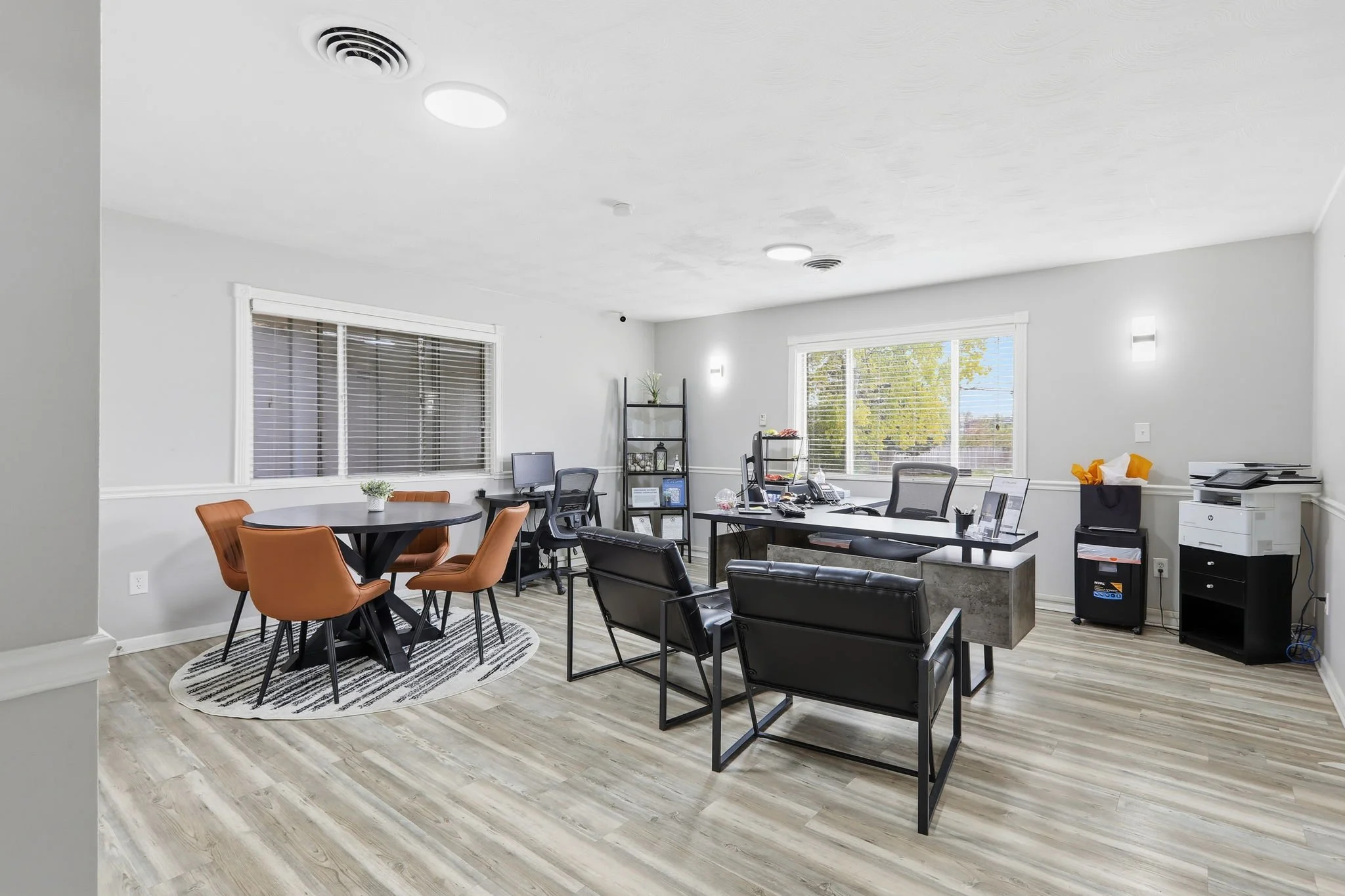 An office space with multiple workstations, chairs, a round table with orange chairs, a black chair, a black shelving unit, and a copier. Large windows with blinds let in natural light. The ceiling has several lights and vents. Light wood flooring.
