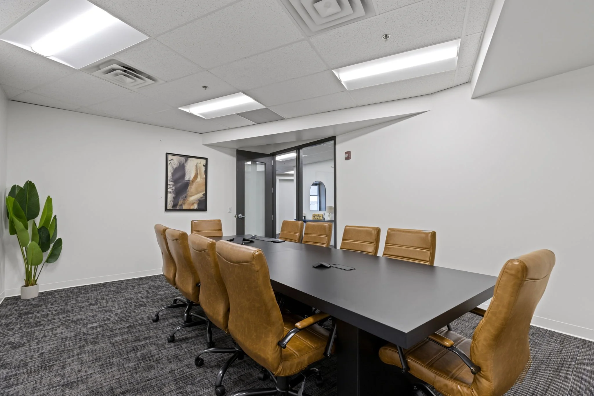 A modern conference room with a long black table surrounded by tan leather chairs. The room has white walls, a framed abstract art piece, a green plant, and a glass door. The ceiling has white panels and fluorescent lighting.