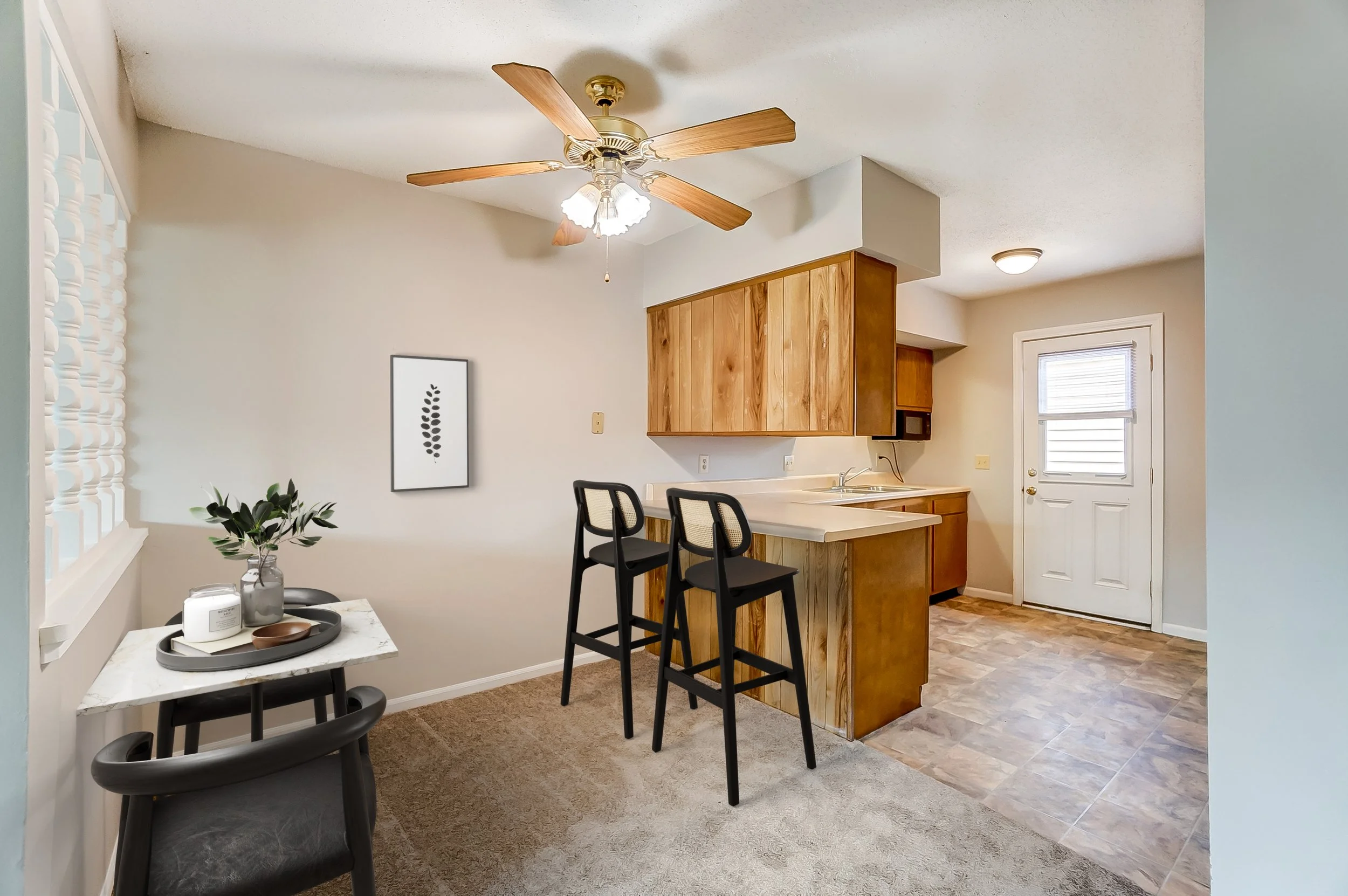A small dining area with a white marble table and black chairs, a decorative plant in a glass jar, on the wall a framed black leaf artwork, adjacent to a small kitchen with wooden cabinets, a white countertop, two black barstools, a ceiling fan with 