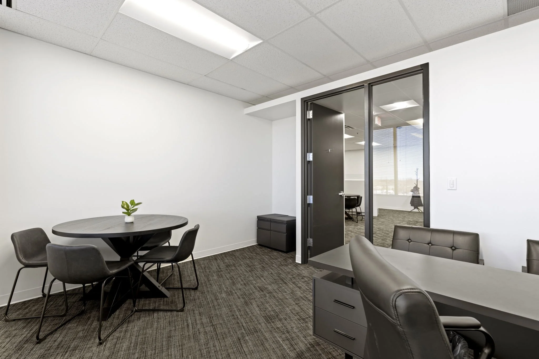 Empty modern office meeting room with a round table, black chairs, a plant, and a black cabinet, with a glass door leading to another room with chairs and large windows.