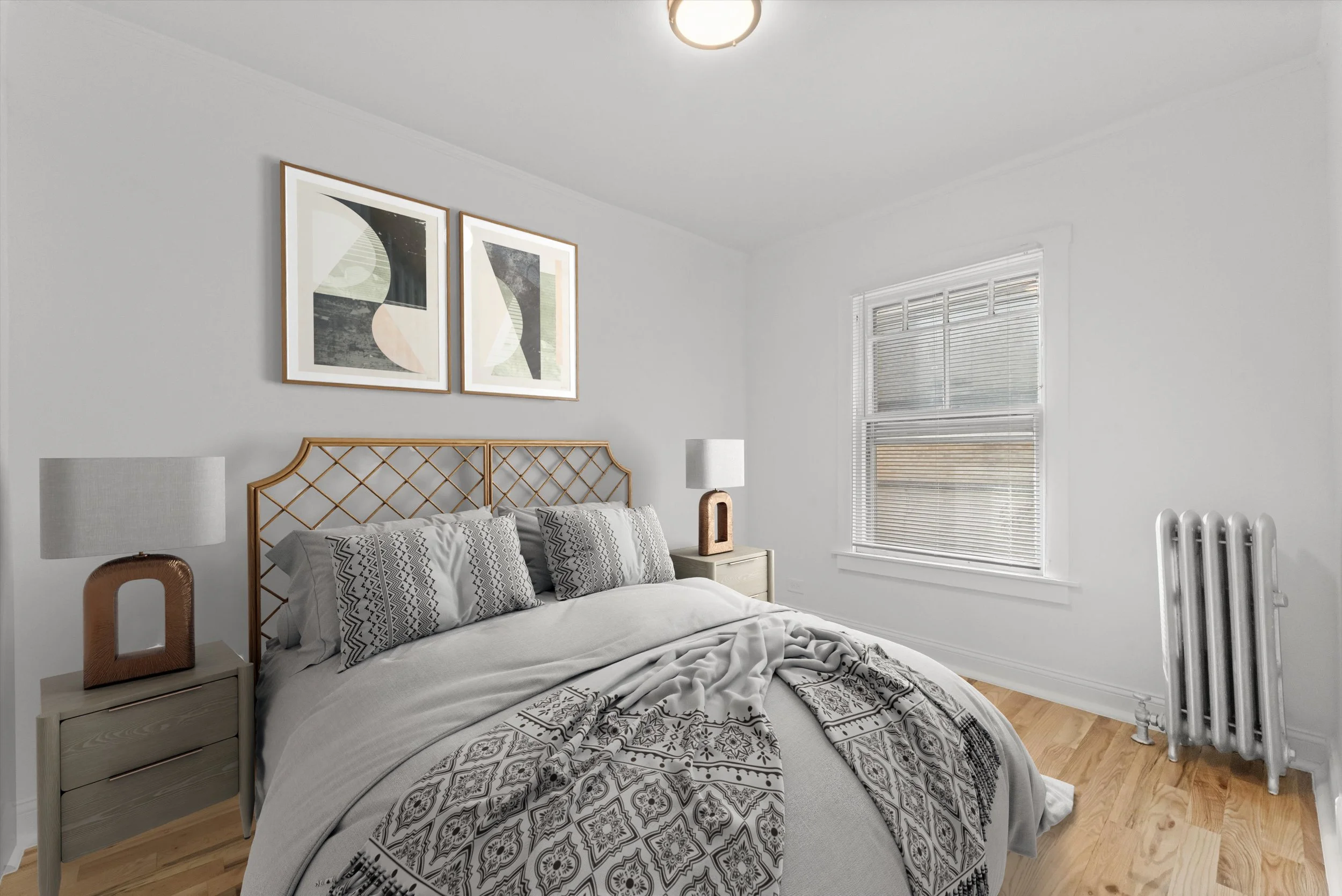 A bedroom with white walls, a bed with patterned bedding, a wooden headboard, and two matching bedside tables with white lamps. There are two framed abstract art pieces above the bed and a window with blinds to the right.