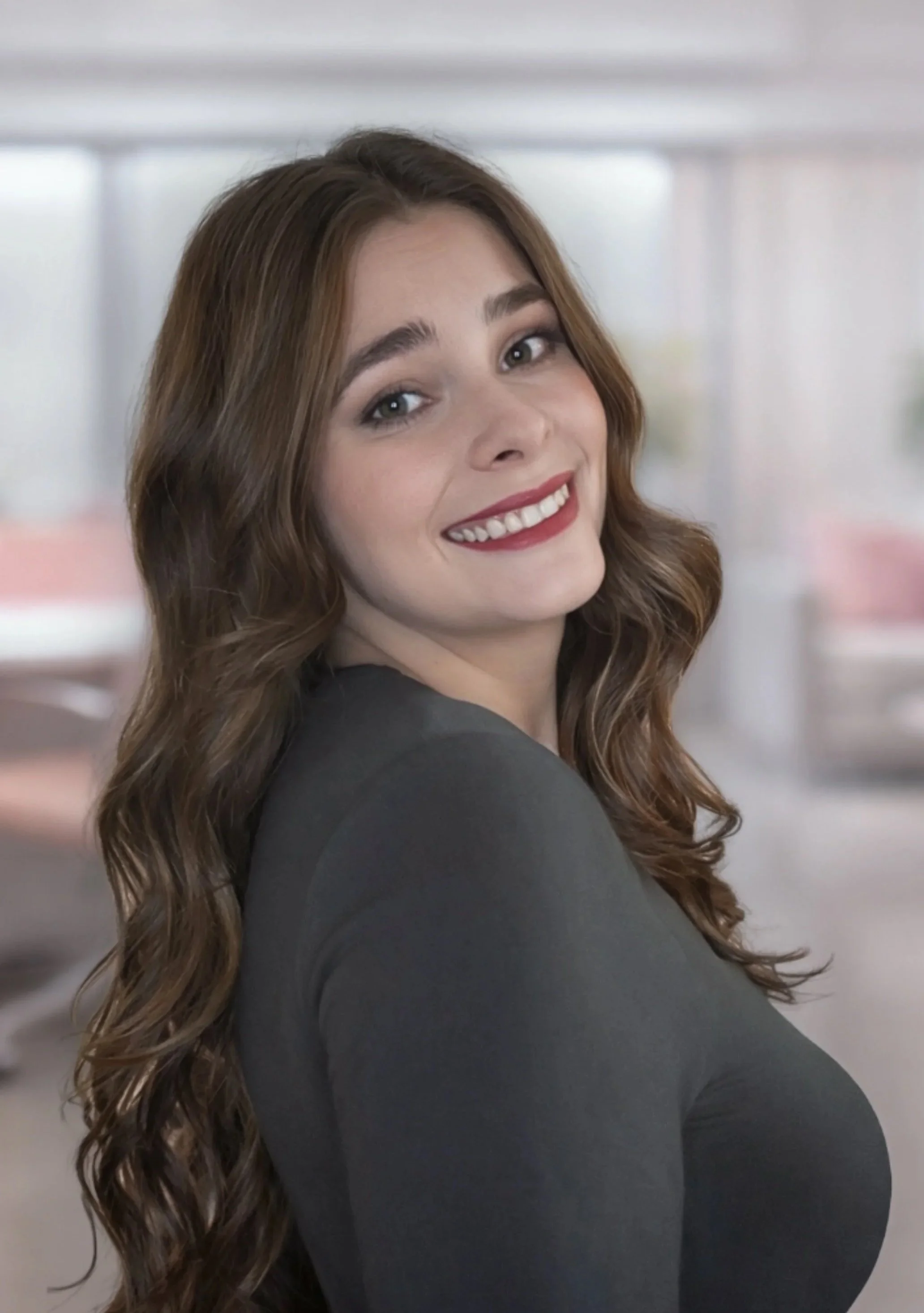 A woman with long, wavy brown hair and fair skin smiling at the camera, wearing a dark top, in an indoor setting with a blurred background.