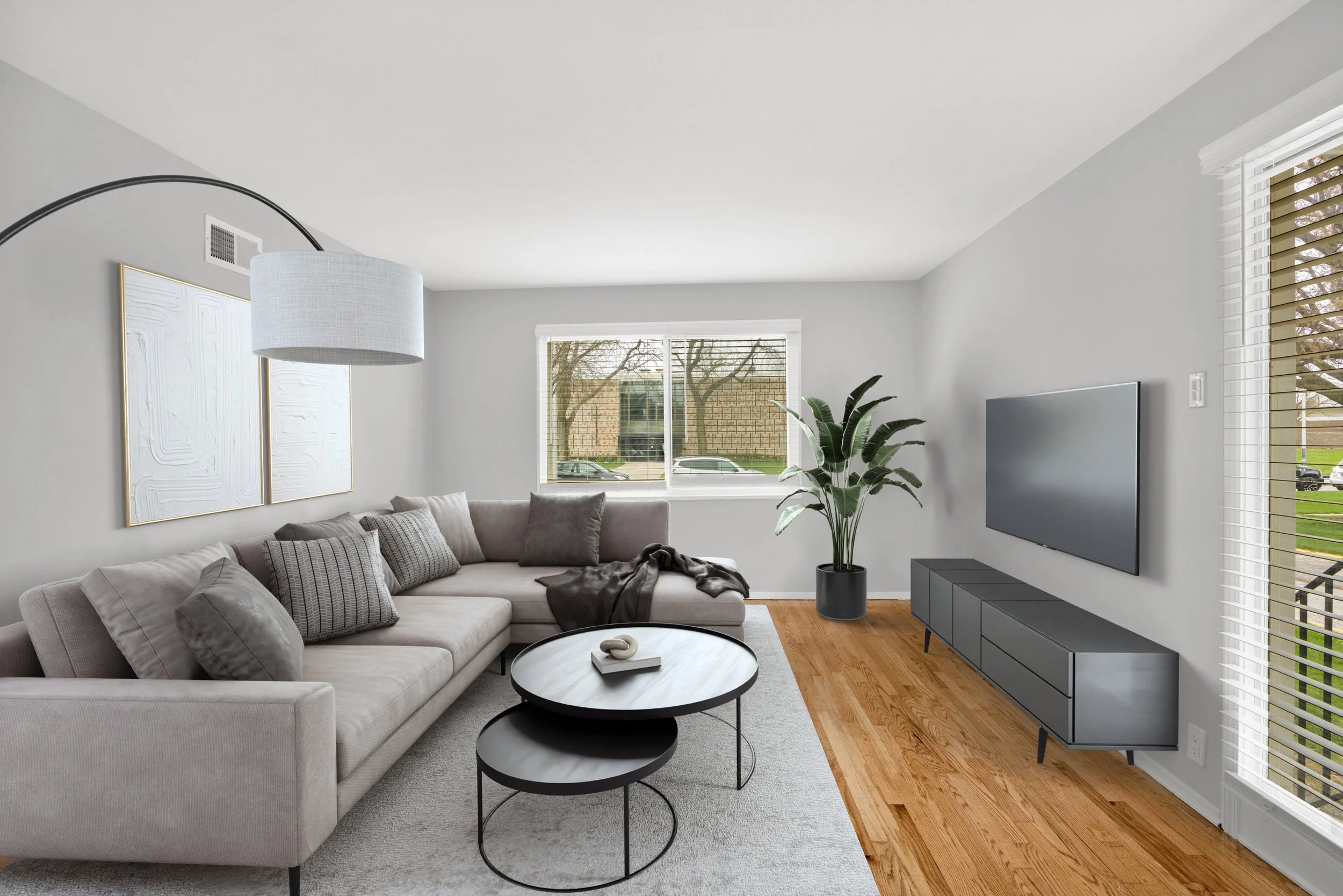Living room with beige sectional sofa, black round coffee tables, wall-mounted flat-screen TV, large windows, hardwood floors, and a potted plant.