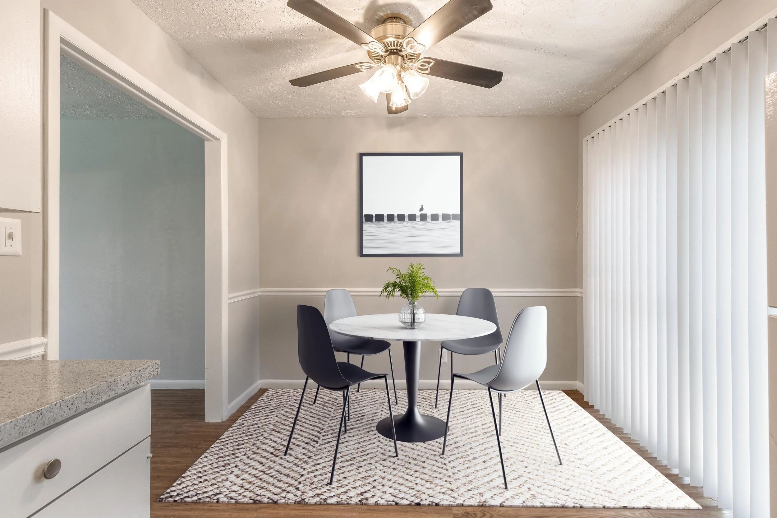 A dining area with a round black table surrounded by black and white chairs, a window with vertical blinds, a ceiling fan with lights, a picture on the wall, and a plant on the table, with a light-colored rug on a hardwood floor.