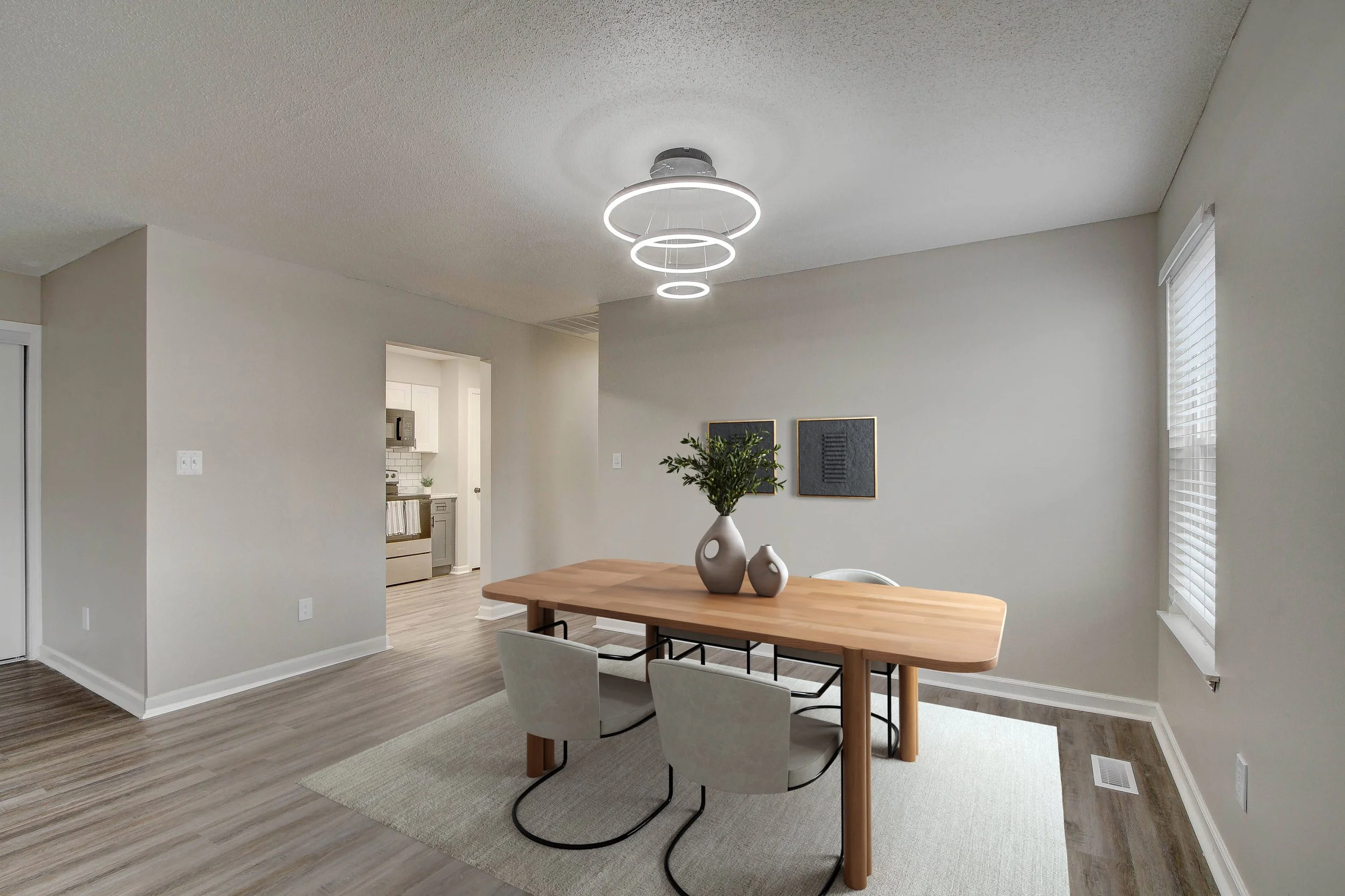 A modern dining room with a wooden table, white chairs, a light fixture with three illuminated rings hanging from the ceiling, a potted plant, and wall art, with a view into the kitchen area.