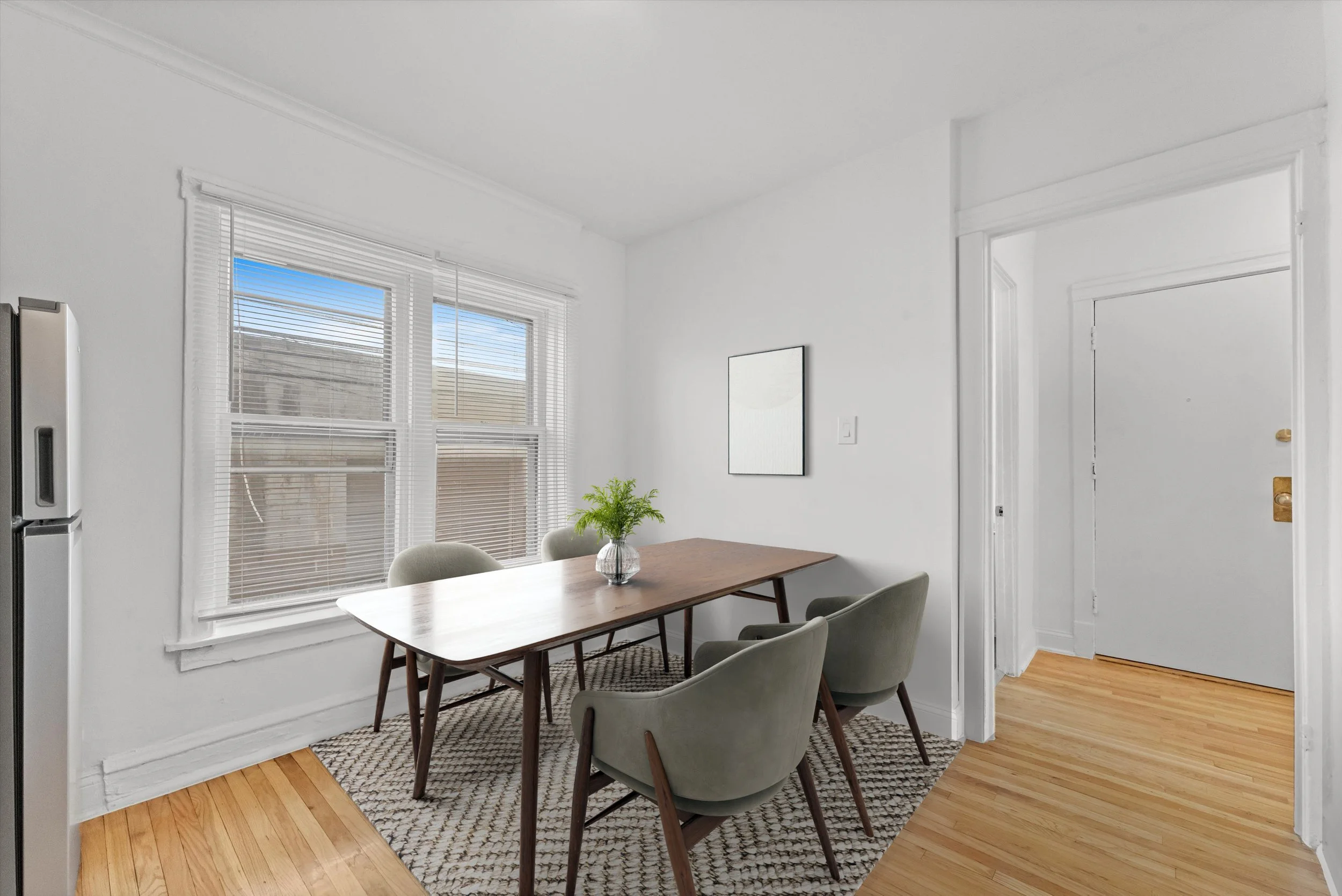 A dining room with a wooden table, four upholstered chairs, large windows with horizontal blinds, a small plant in a vase, and a wall mirror, with hardwood flooring and white walls.