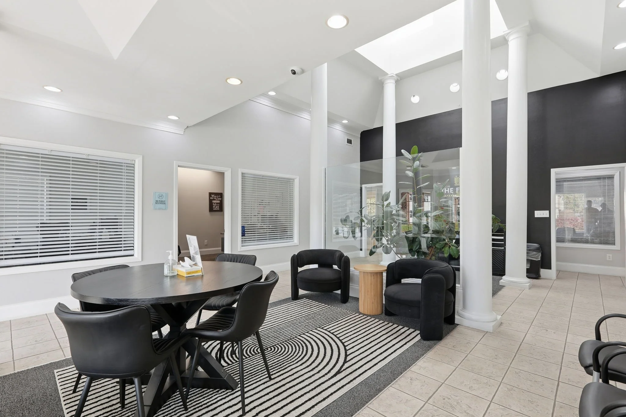 Interior of a modern waiting room with black chairs around a round table, white walls, multiple windows with blinds, and columns, with a black accent wall and indoor plants.