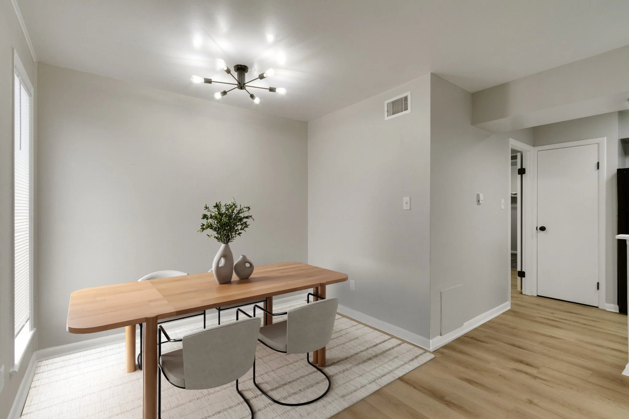 A modern dining room with a wooden table, four gray chairs, a small beige and brown rug, a white vase with green leaves, large windows with blinds, a white door, and a contemporary ceiling light fixture.