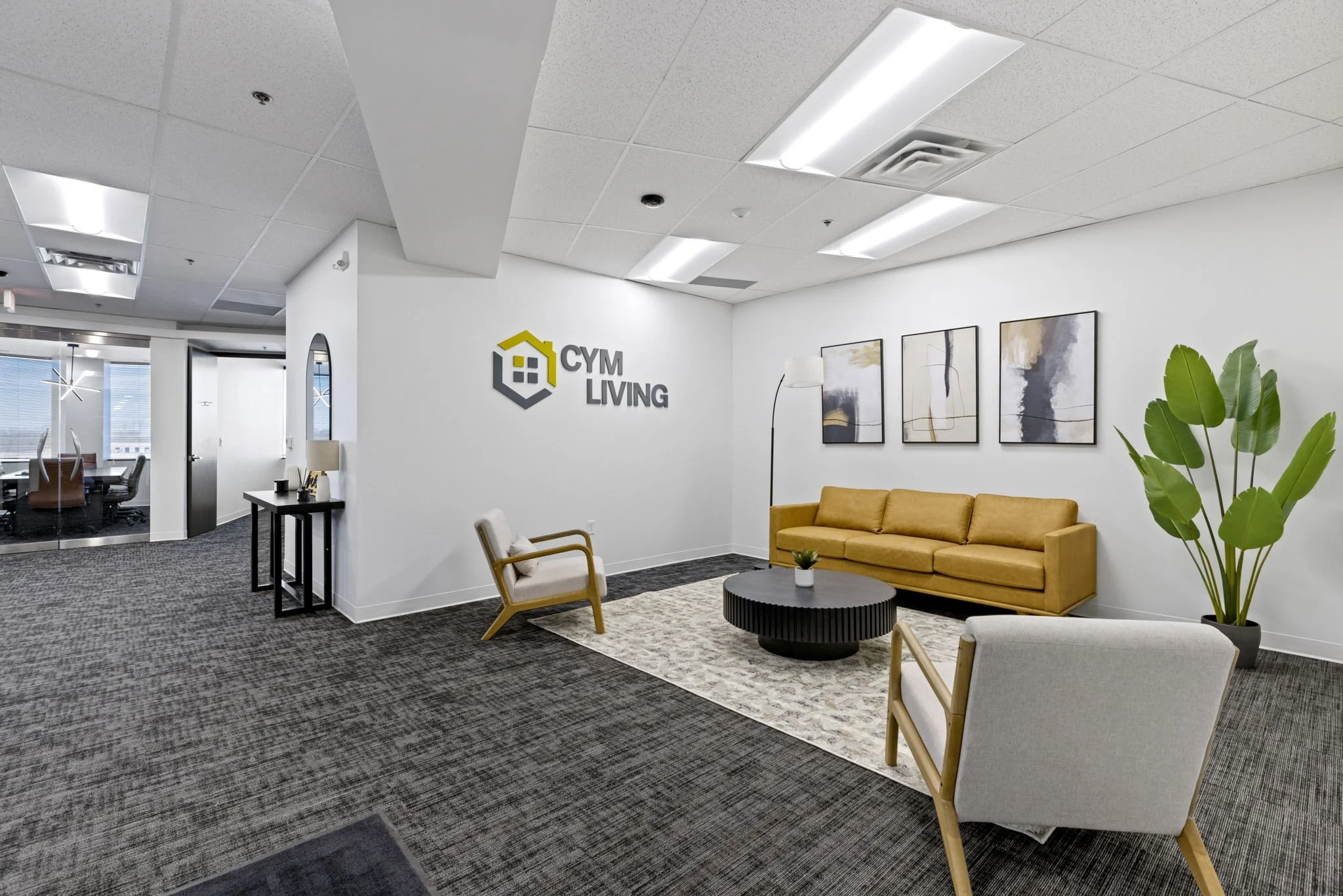 View of an office waiting area with modern decor, including a yellow sofa, armchairs, abstract artwork, a large plant, and the company logo 'CYM Living' on the wall.