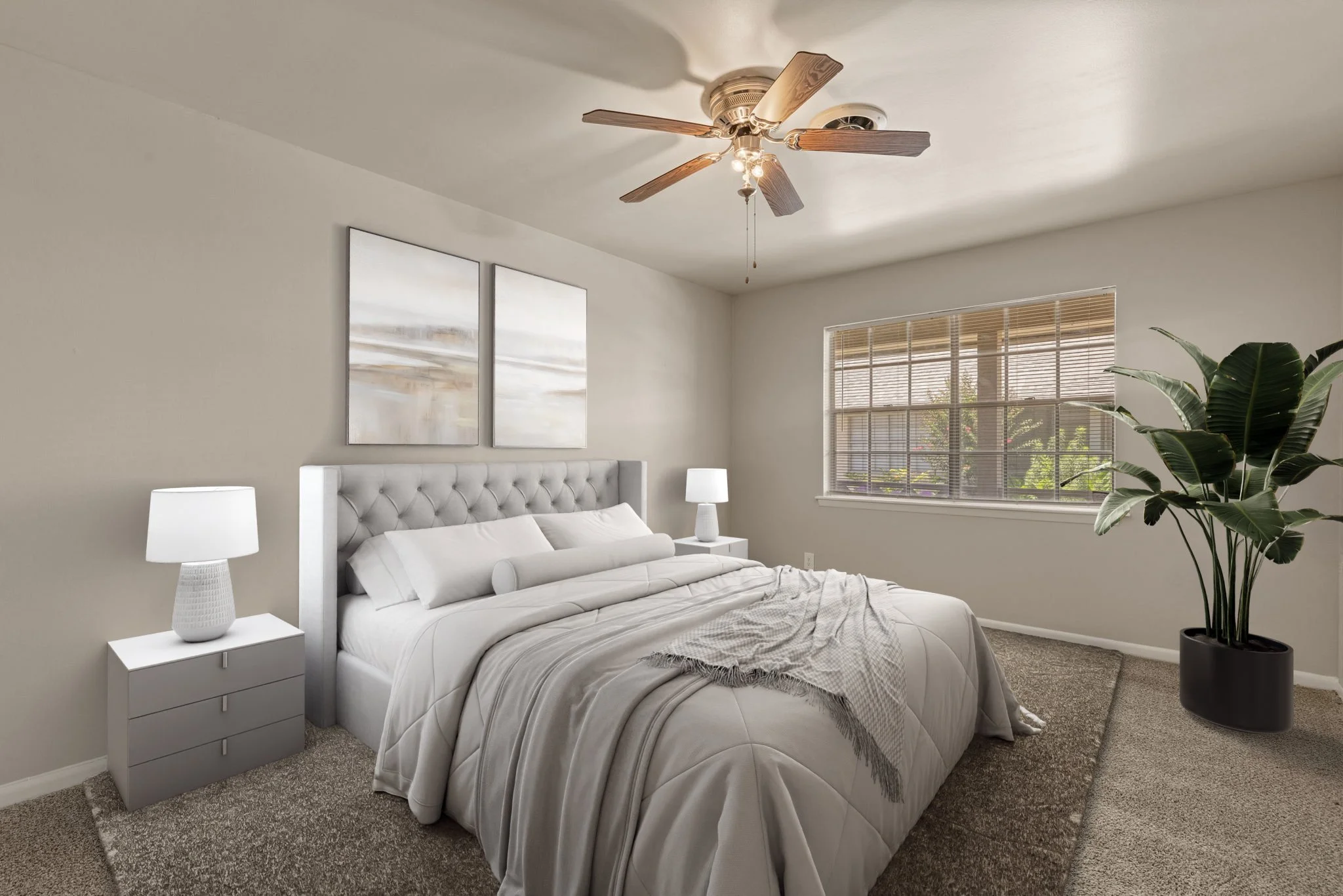 A bedroom with a white tufted bed, nightstands with white lamps, beige walls, and a large window with blinds. There is a ceiling fan with wooden blades and a large potted plant in the corner.
