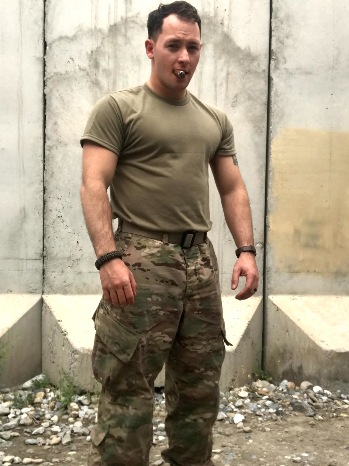 A young man in military camouflage pants and a khaki t-shirt standing outdoors against a concrete wall background.