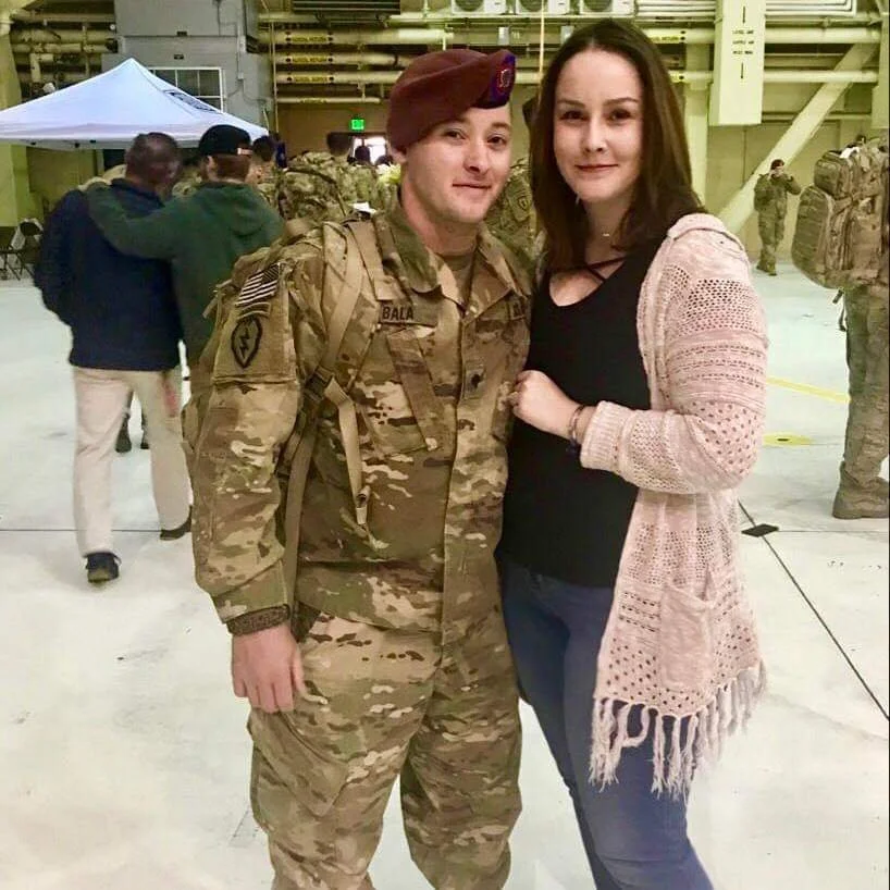 A person in military uniform standing next to a woman in casual clothing inside a military hangar, with other soldiers and civilians in the background.