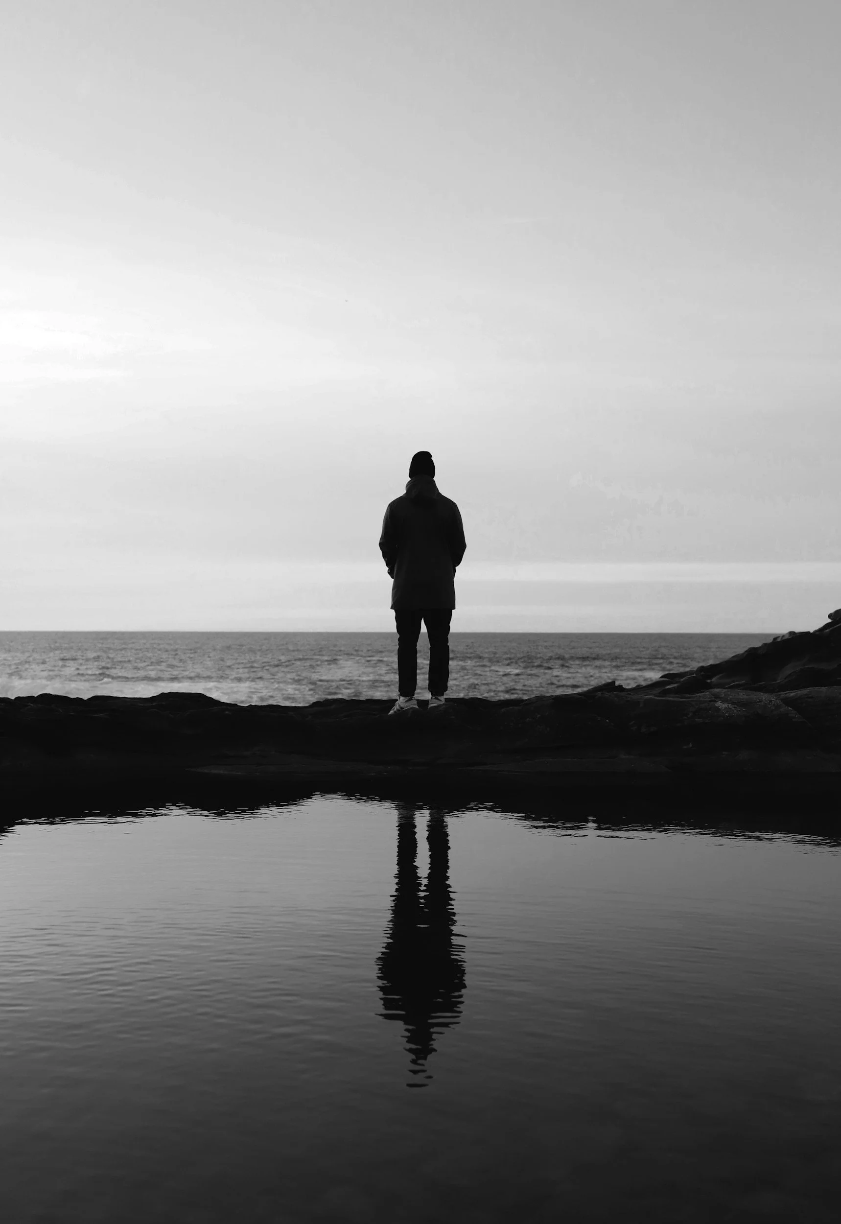 A person standing on rocks near water, looking out at the ocean or sea under an overcast sky, with their reflection visible in the water.