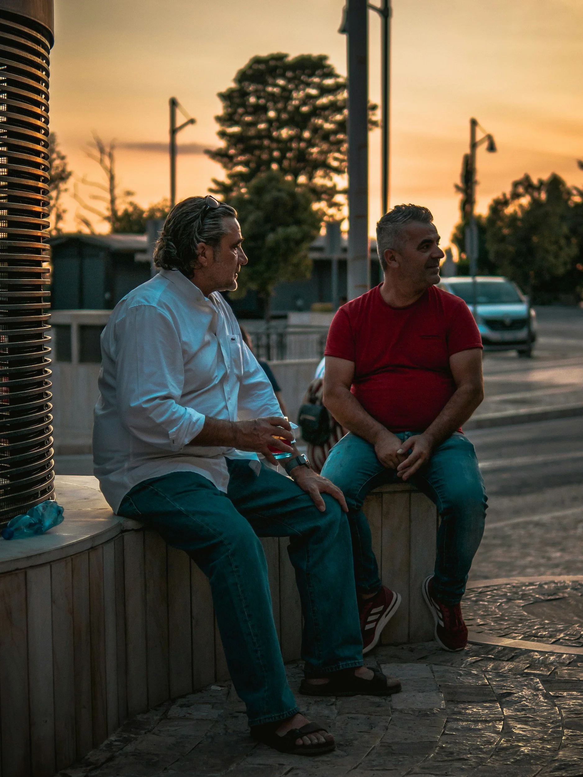 Two men sitting on a wooden bench outdoors during sunset, engaged in conversation, with trees, streetlights, and a parked vehicle in the background.