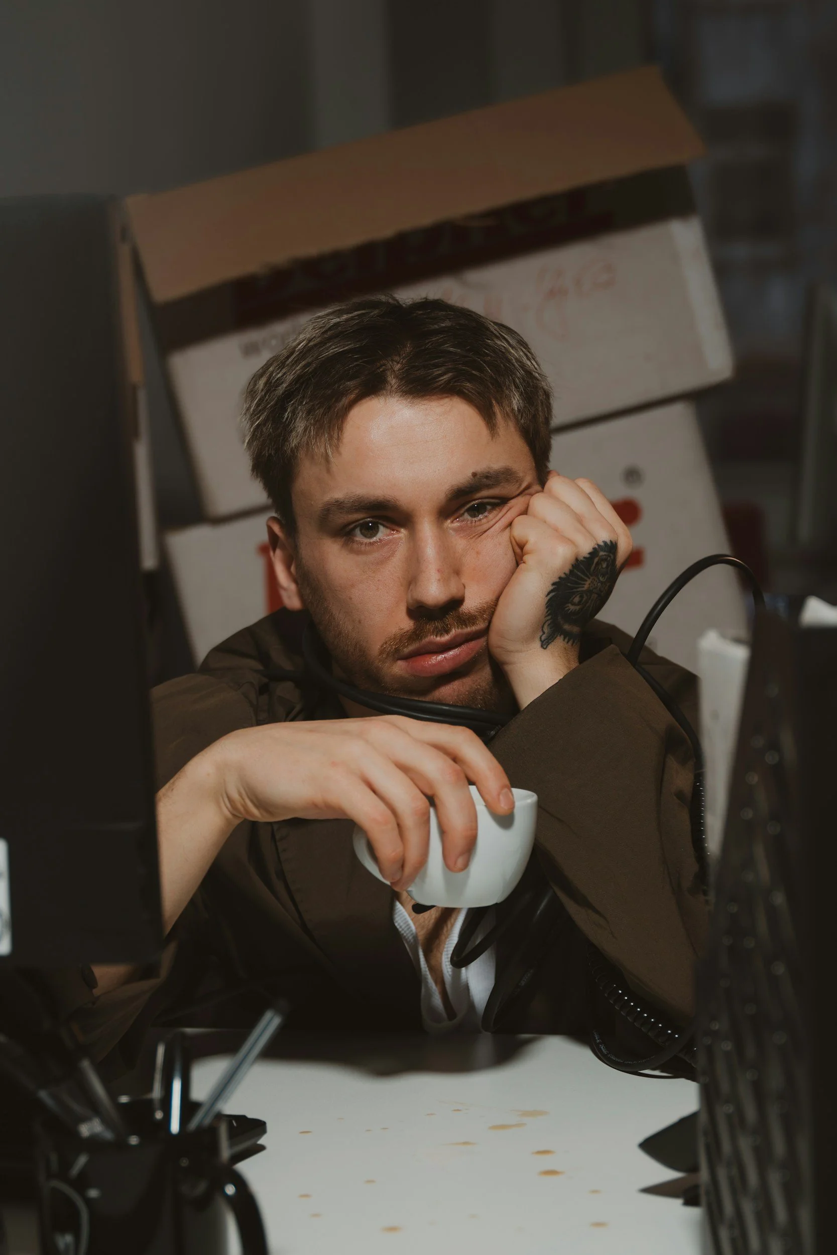 A young man with short brown hair and tattoos on his hand, sitting at a desk with electronics and boxed items in the background, resting his face on his hand and looking at the camera, holding a white mug.