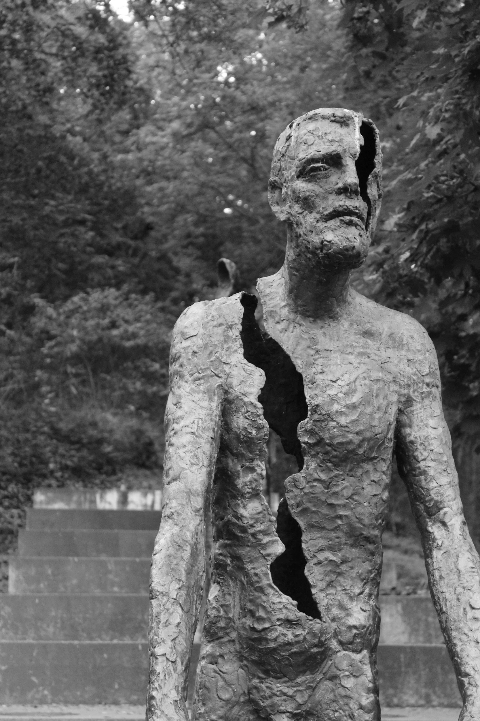 A black and white photo of a damaged and weathered statue of a human figure, with a hollowed-out section exposing the interior, set outdoors with trees in the background.
